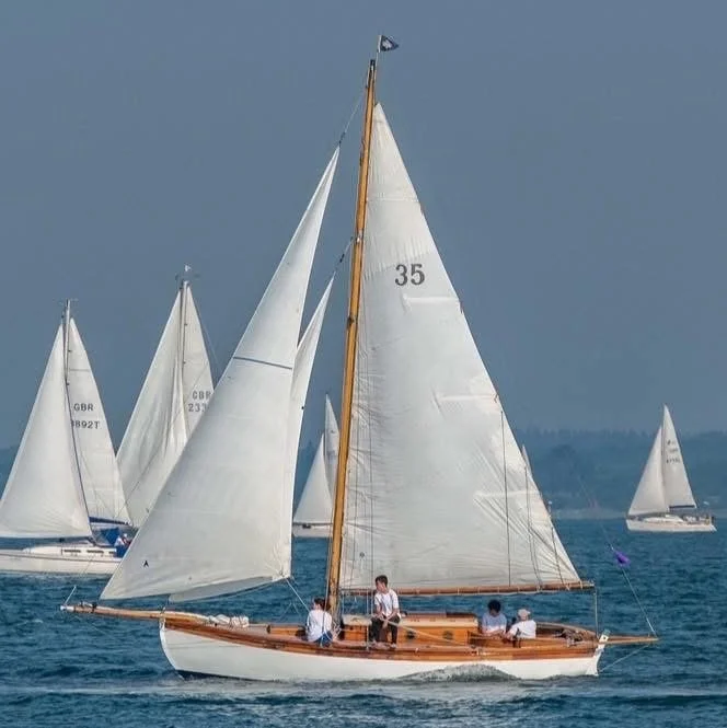 Sailboats on the water during a sailing race, with a focus on a classic wooden boat and several modern white sailboats in the background.