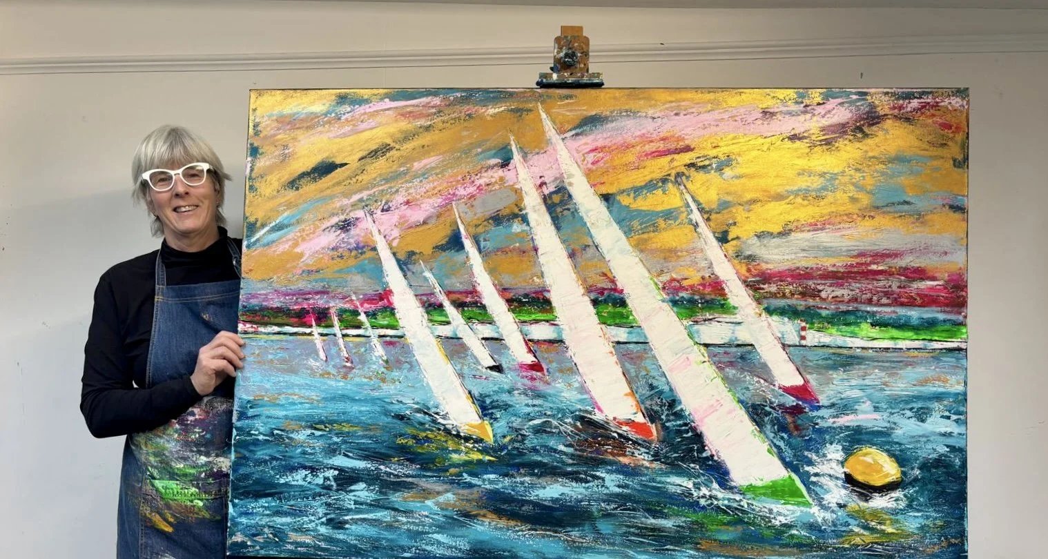 An artist with gray hair, white glasses, and a black shirt, wearing a colorful apron, holding a large abstract painting of sailboats on water with a vibrant, colorful sky in the background.