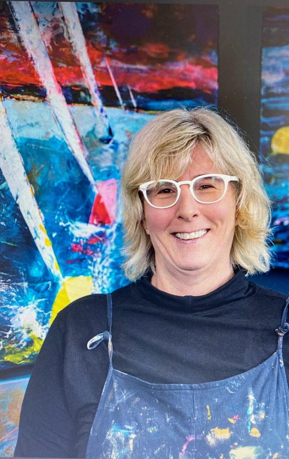 A smiling woman with blond hair, wearing white glasses and a black top, standing in front of a colorful abstract painting.