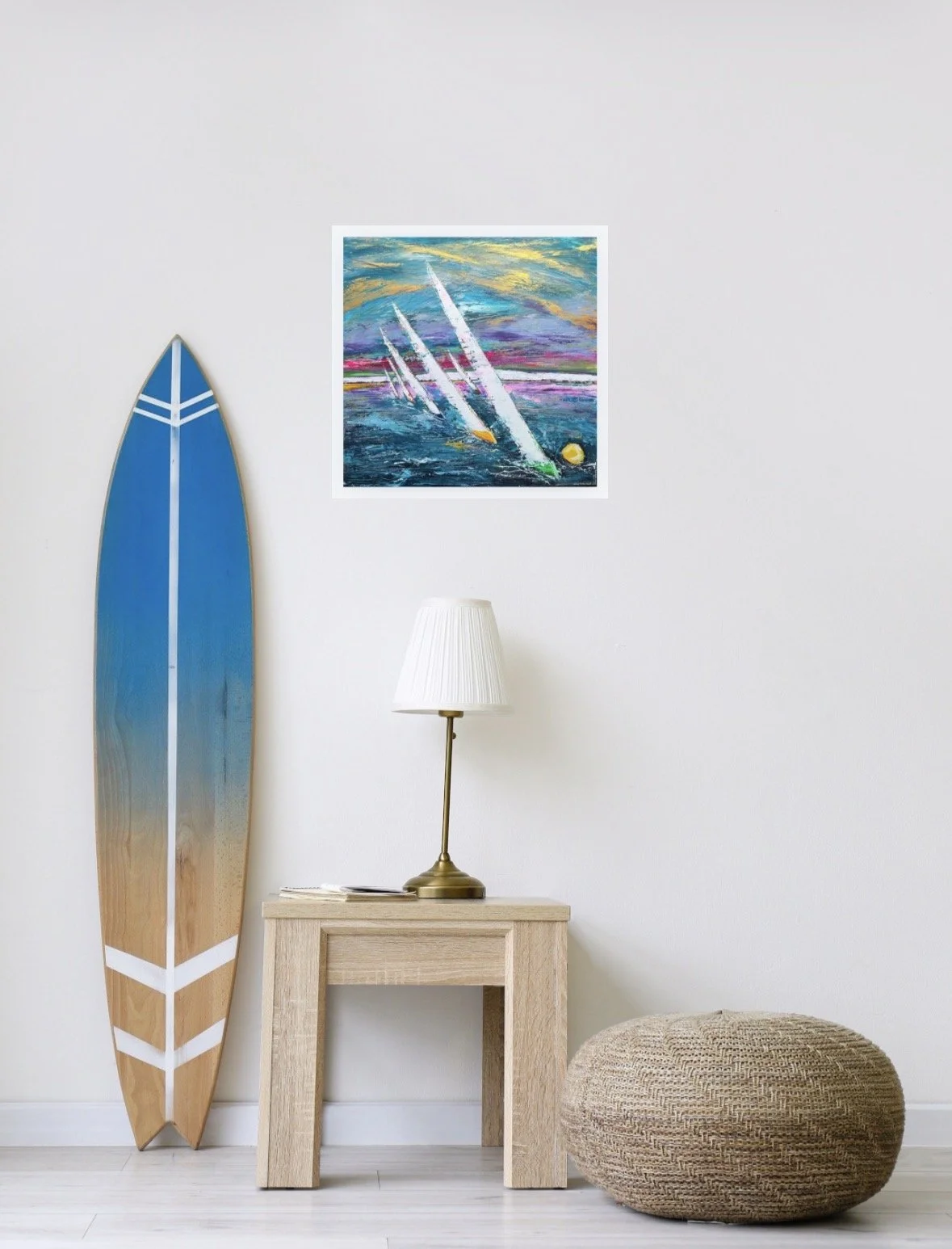 A surfboard leaning against a white wall, a small wooden side table with a white lamp and some books, a round woven pouf, and a colorful abstract painting hanging on the wall.