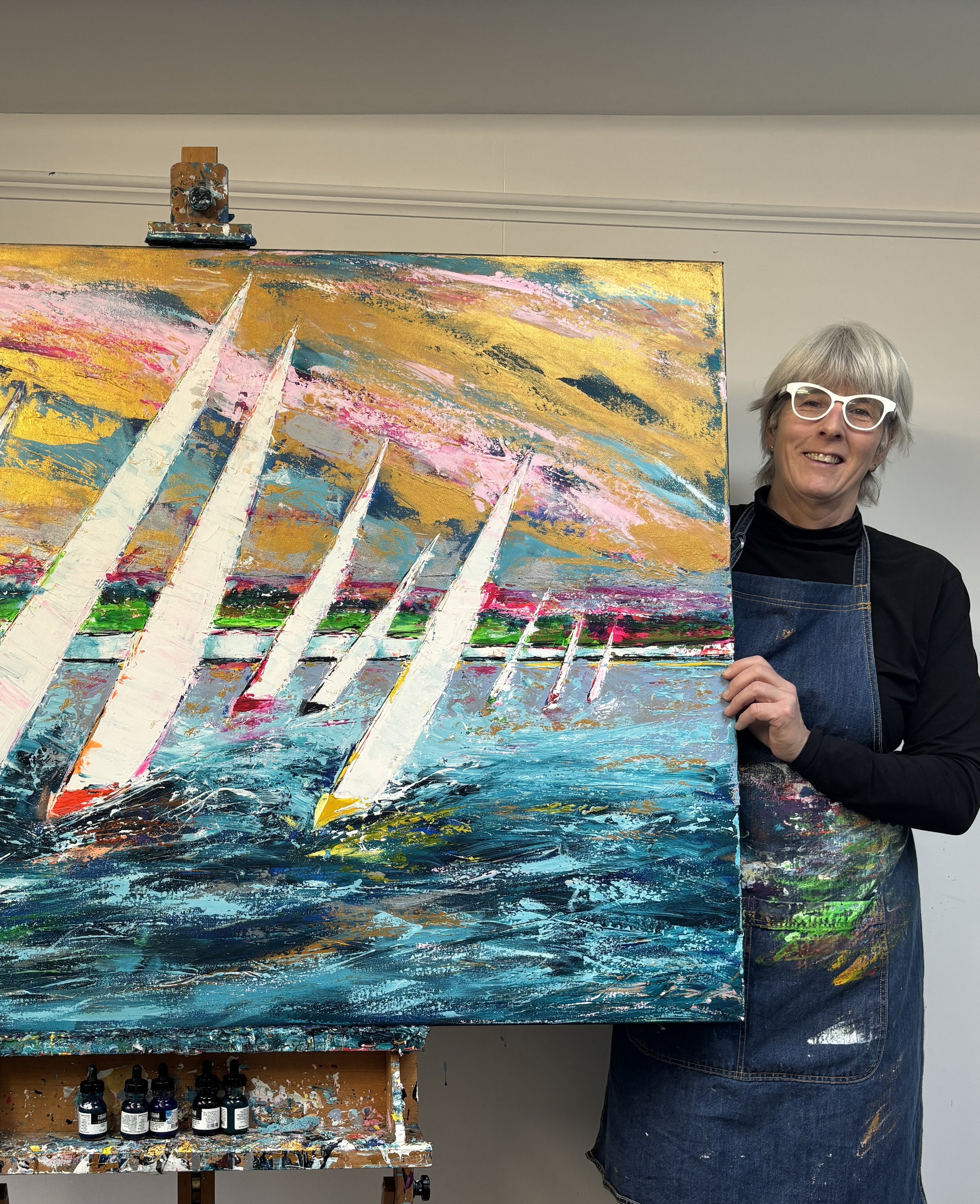 A woman with short gray hair and white glasses holding an abstract painting of sailboats on water with a colorful sky behind them.