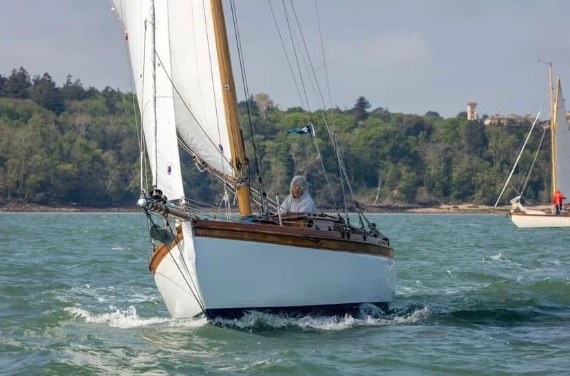 A sailboat on the water with a person on board, sailing near a wooded coastline with hills and a lighthouse in the background.