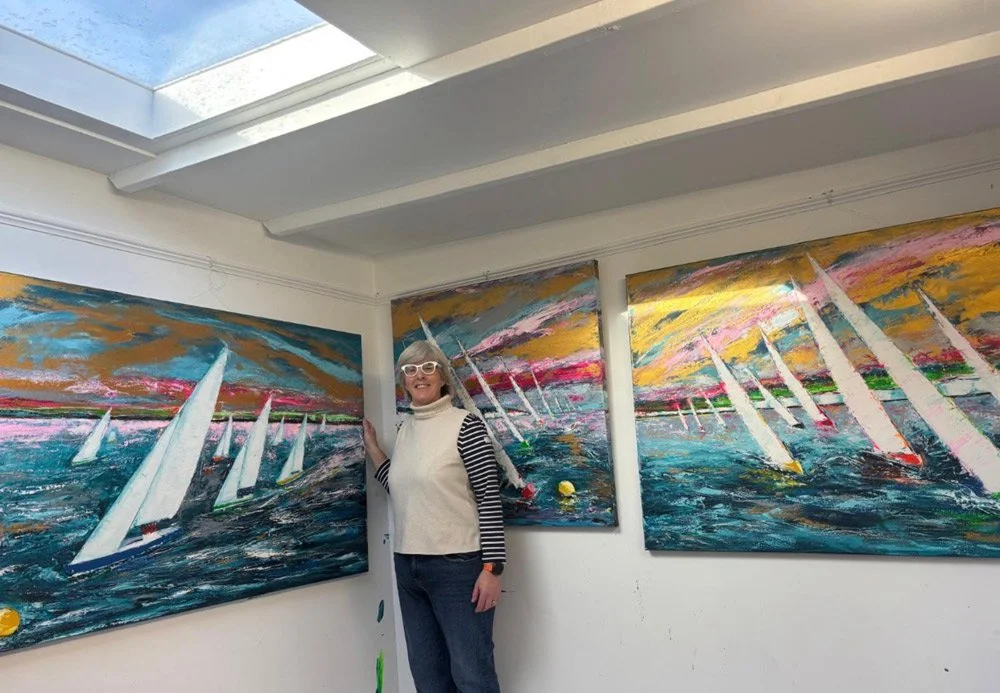 A woman with white glasses and a striped long sleeve shirt standing in front of three paintings of sailboats on water, with a skylight on the ceiling.