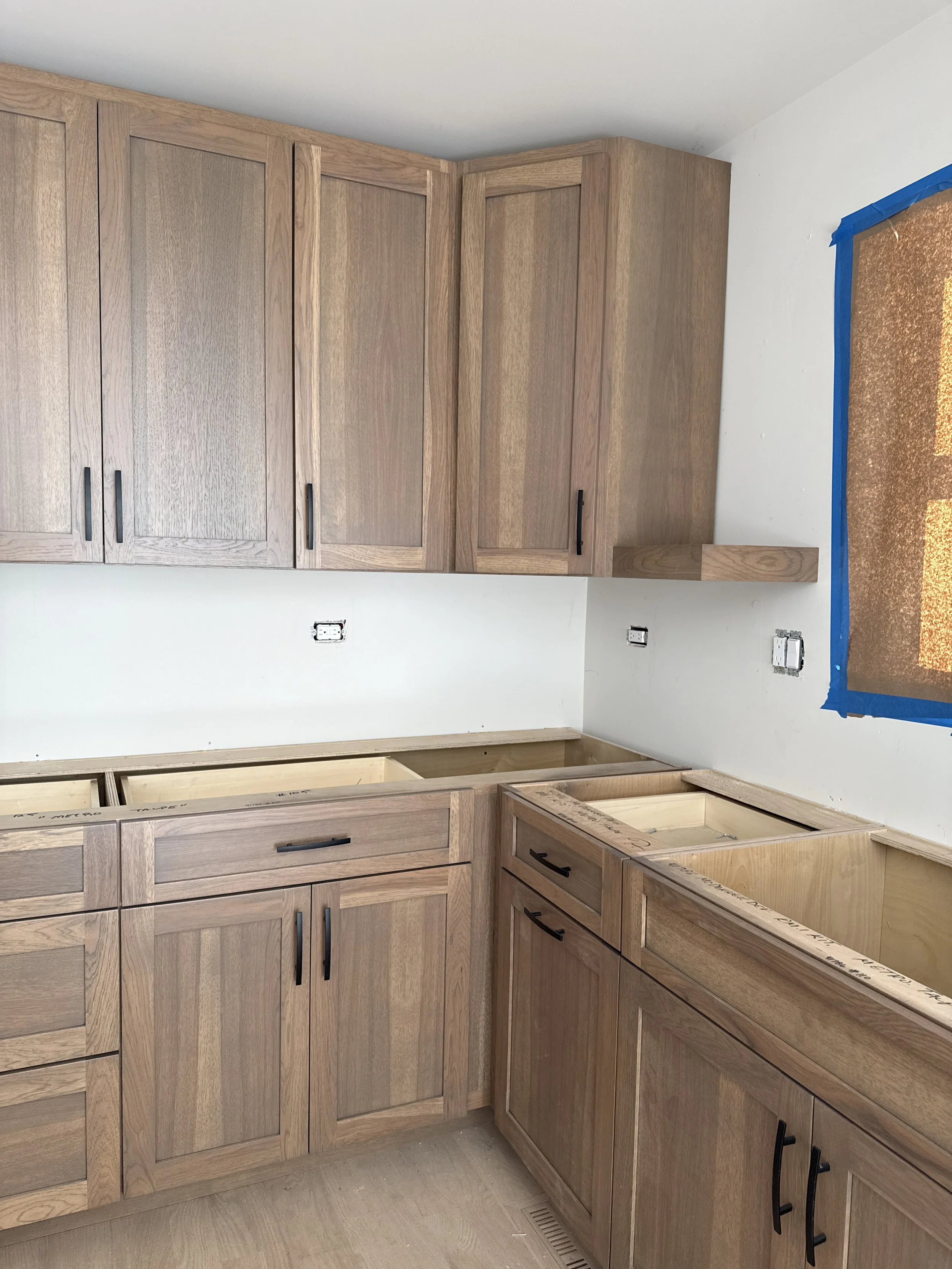 Kitchen with wooden cabinets, some drawers and cabinet doors installed, with unfinished countertop space, and a window with blue painter's tape around it, indicating ongoing construction.