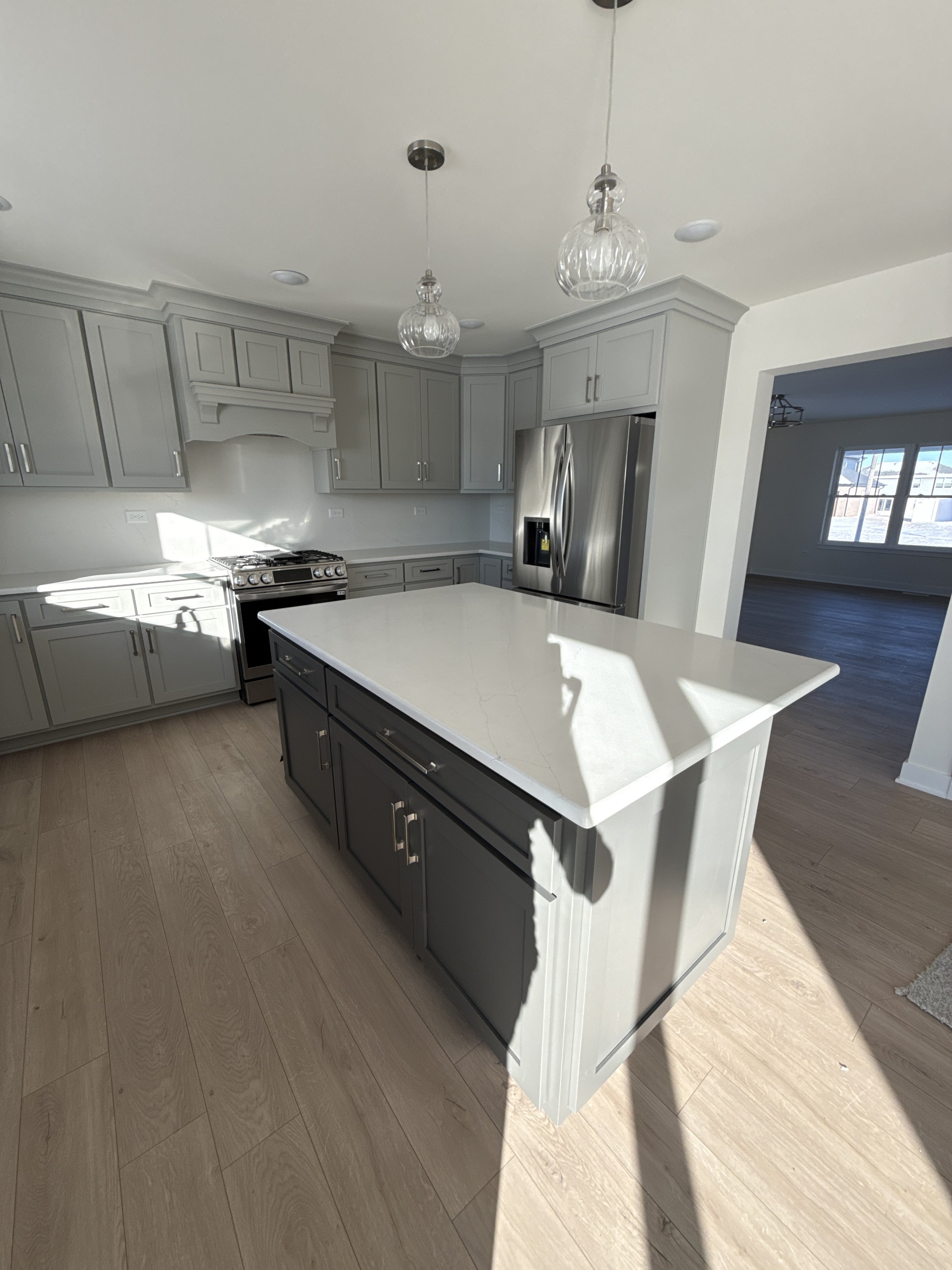 A modern kitchen with light gray cabinets, a white countertop island, stainless steel refrigerator, oven, and light hardwood floors. Clear glass pendant lights hang from the ceiling, and sunlight illuminates the space.