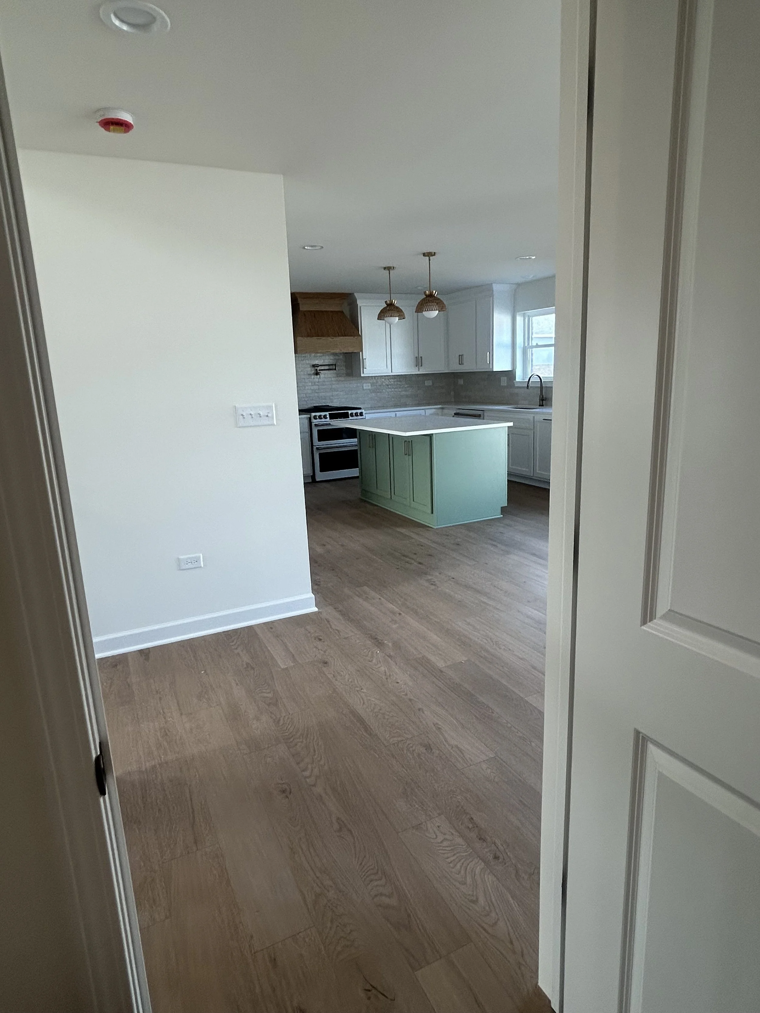 View of a modern kitchen with white cabinets, a light green kitchen island, hardwood floors, pendant lights, and a window over the sink.