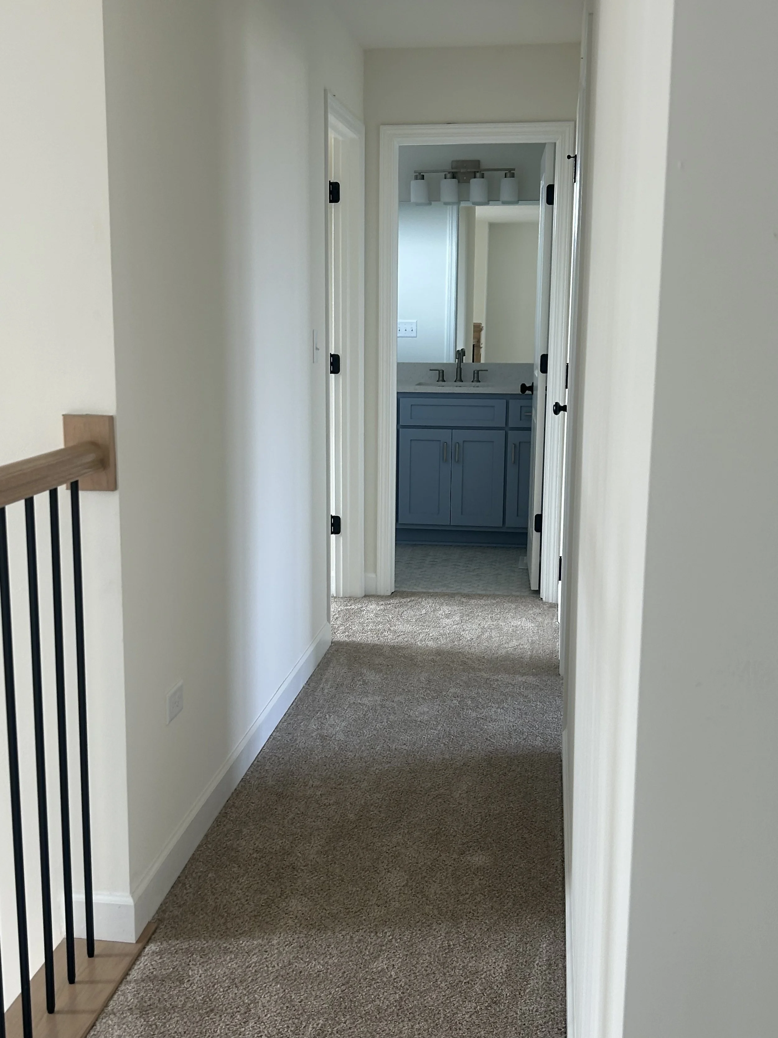 A hallway leading to a bathroom with a blue vanity and a mirror above the sink. The hallway has beige carpet and white walls.