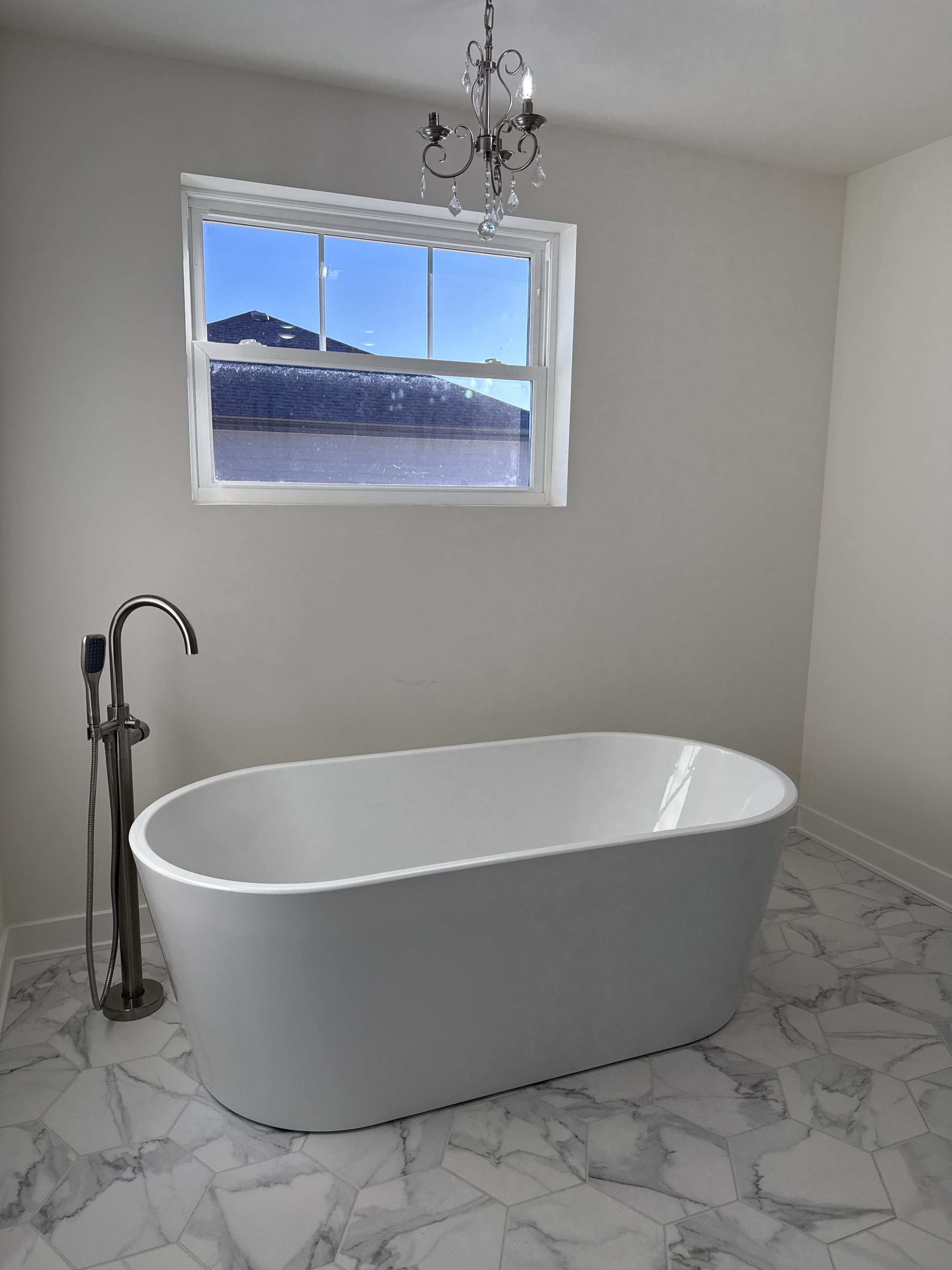 Bathroom with a standalone white bathtub, window, chandelier, and marble tile flooring.