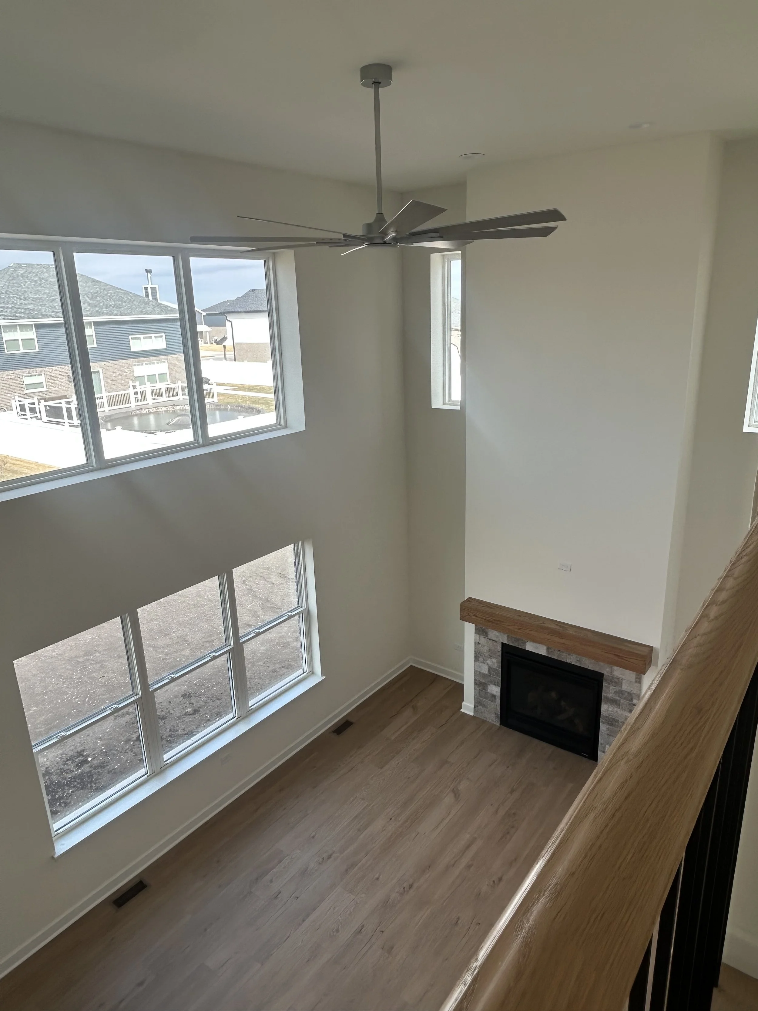 Empty living room with high ceiling, large windows, a fireplace with a stone surround and wooden mantle, and a ceiling fan, viewed from an upper level.
