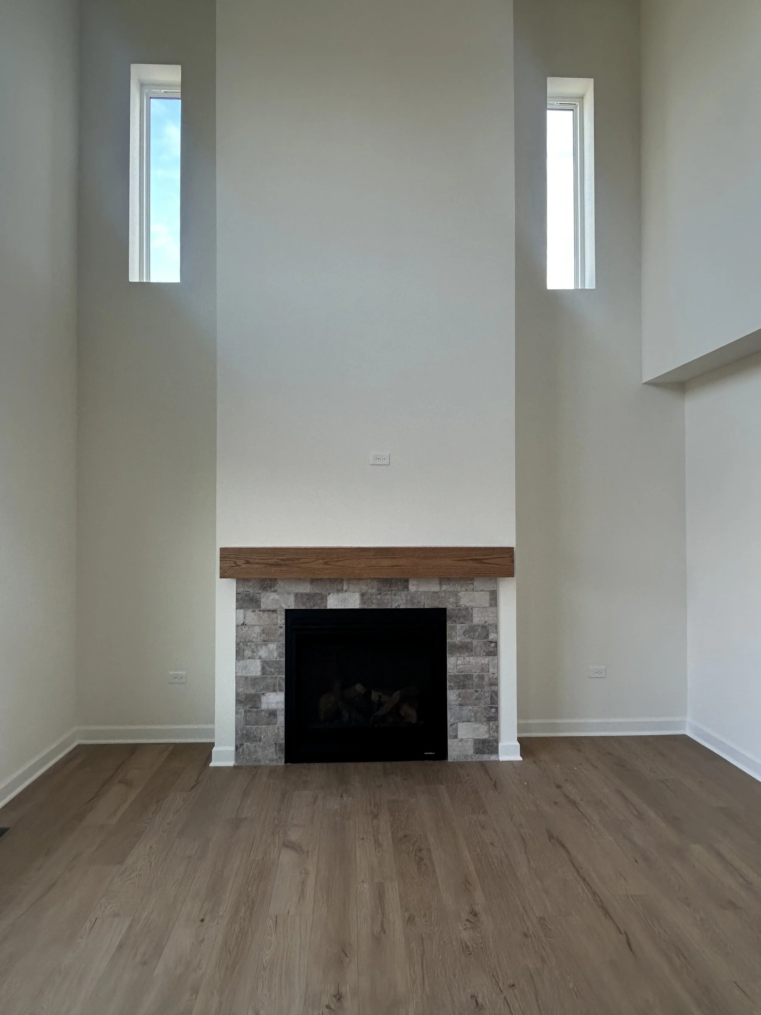 Empty room with a fireplace, two tall narrow windows, beige walls, wooden floor, and minimal decor.