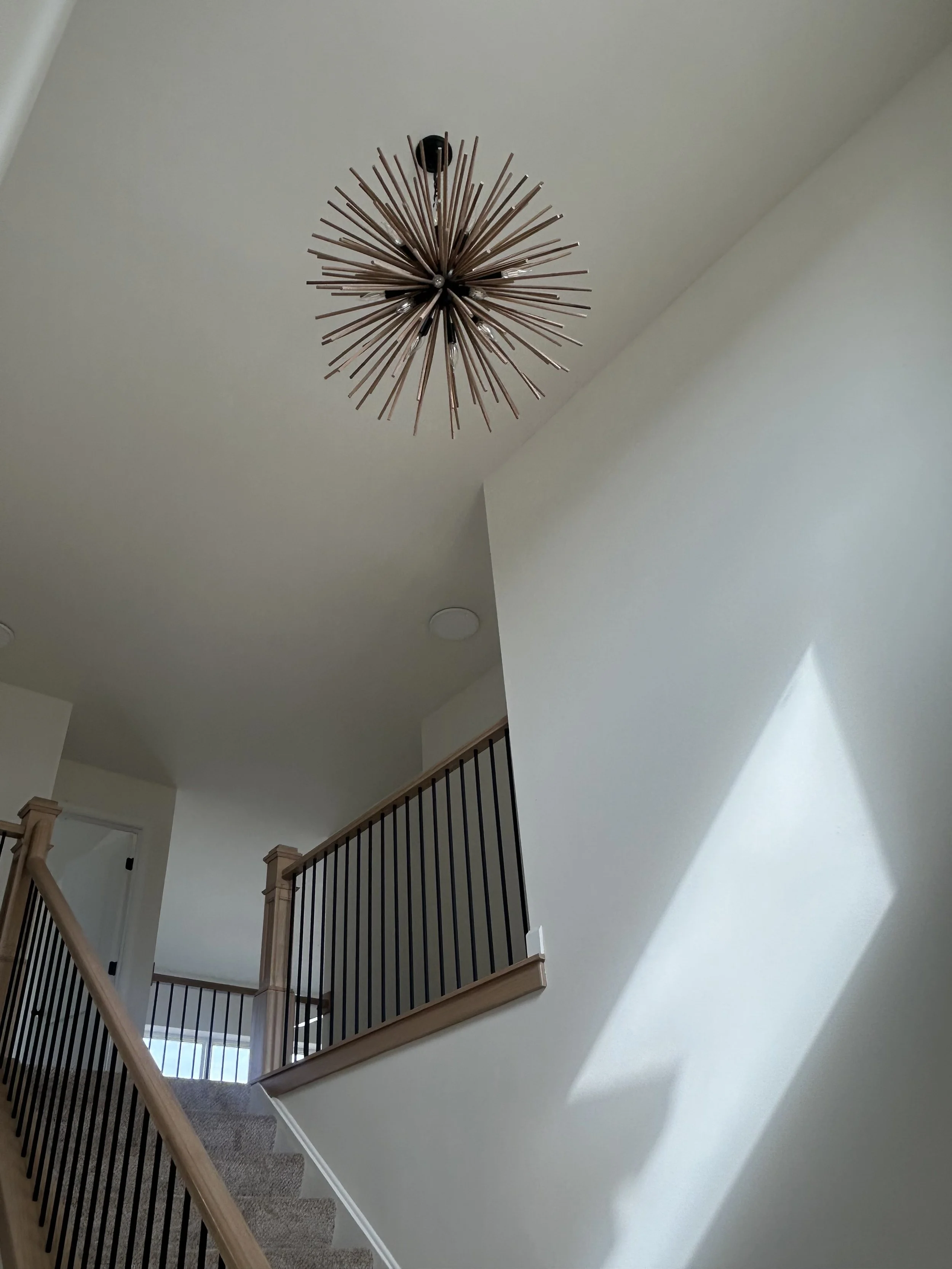 Interior view of a staircase and hallway with a modern chandelier hanging from the ceiling. Sunlight casts a shadow on the wall.