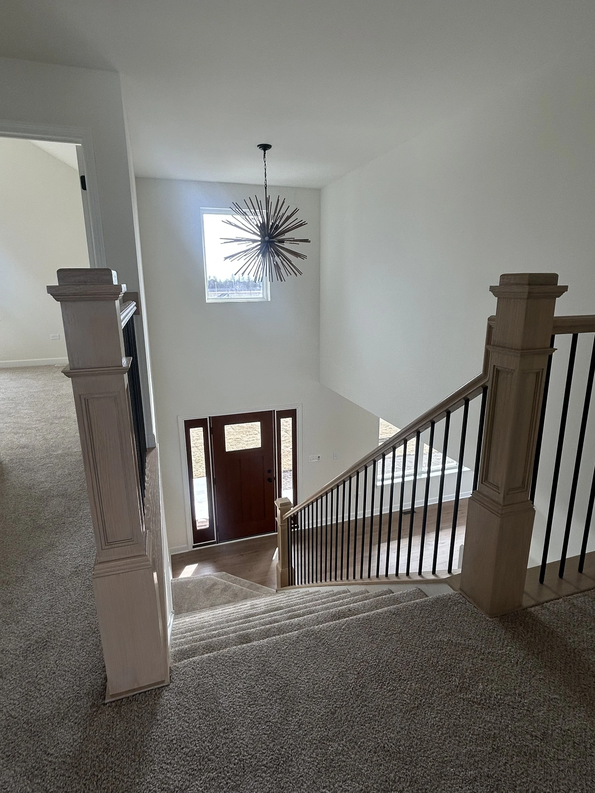 View of a modern home interior with stairway, wooden handrail, front door, window, and a decorative hanging light fixture.