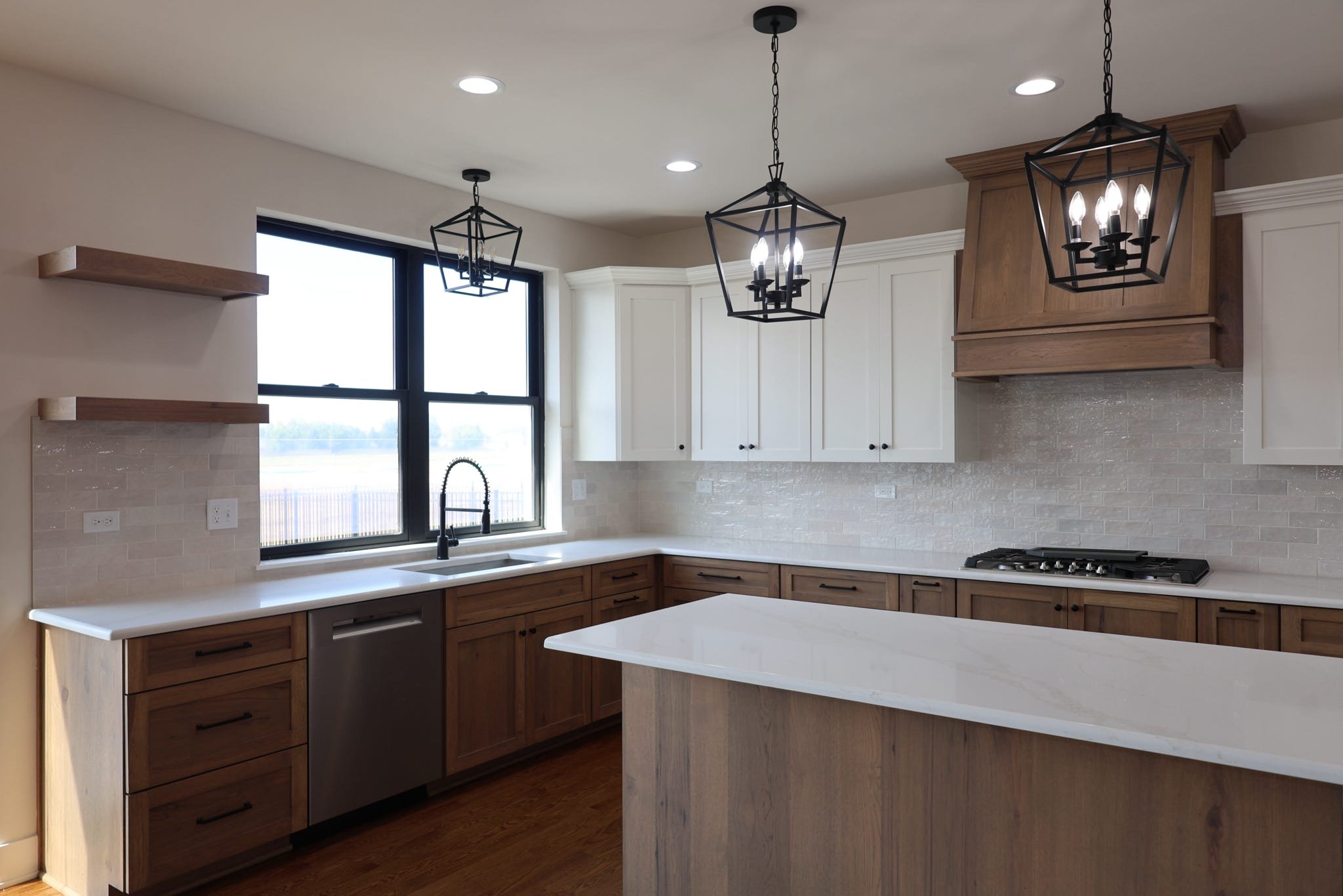 Modern kitchen with white cabinets, wooden lower cabinets, and a white countertop island. There is a large window above the sink, black window frame, and pendant lighting fixtures hanging from the ceiling.