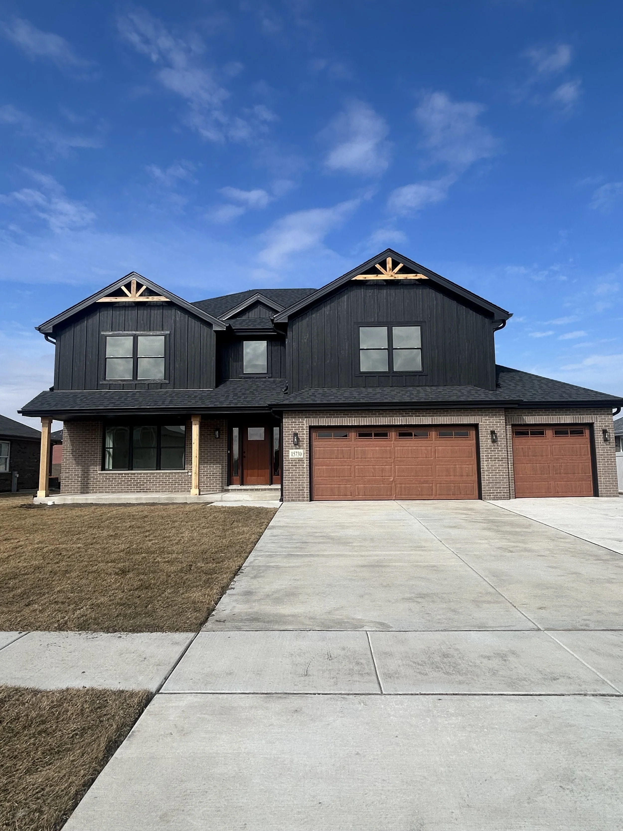 A modern two-story house with black siding and a brick foundation, featuring a front porch supported by wooden posts, large windows, and a three-car garage with wooden doors. The house is set against a blue sky with some clouds, and the front yard ha