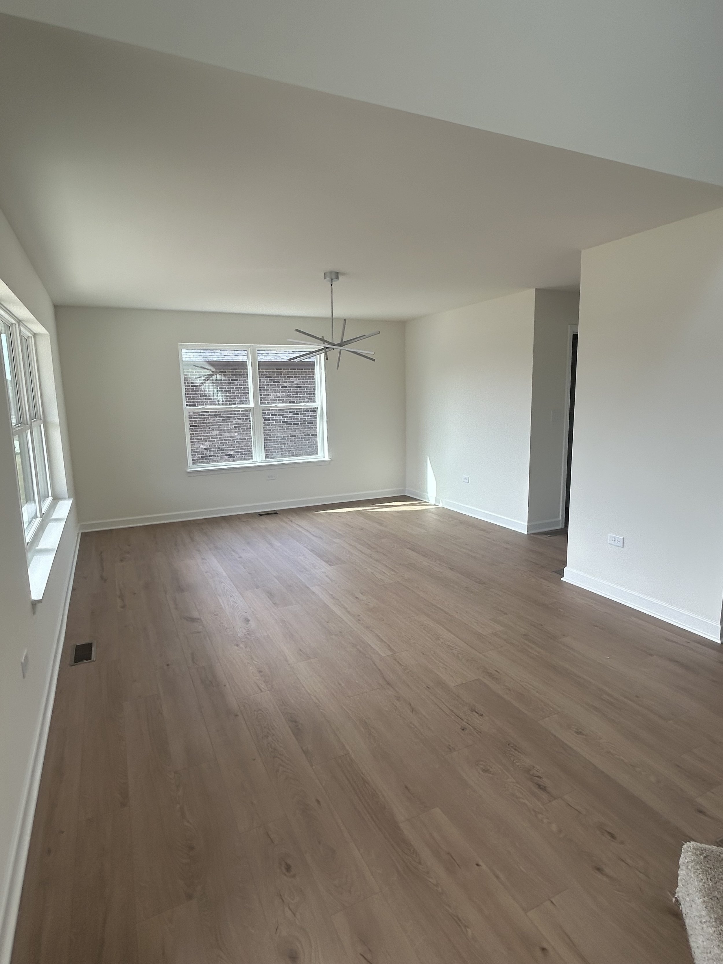 Empty living room with large windows, light-colored hardwood floors, and a modern ceiling fan.