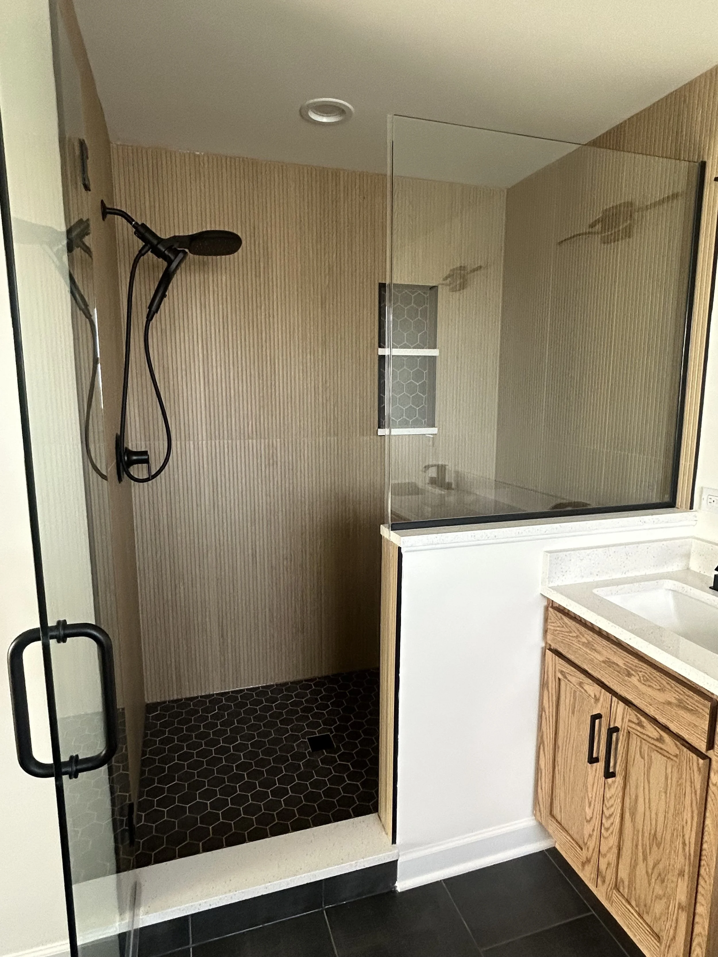 A modern bathroom with a walk-in shower featuring a glass door, black hexagon tiles on the shower floor, a black showerhead, and natural wood cabinetry next to a white countertop sink.