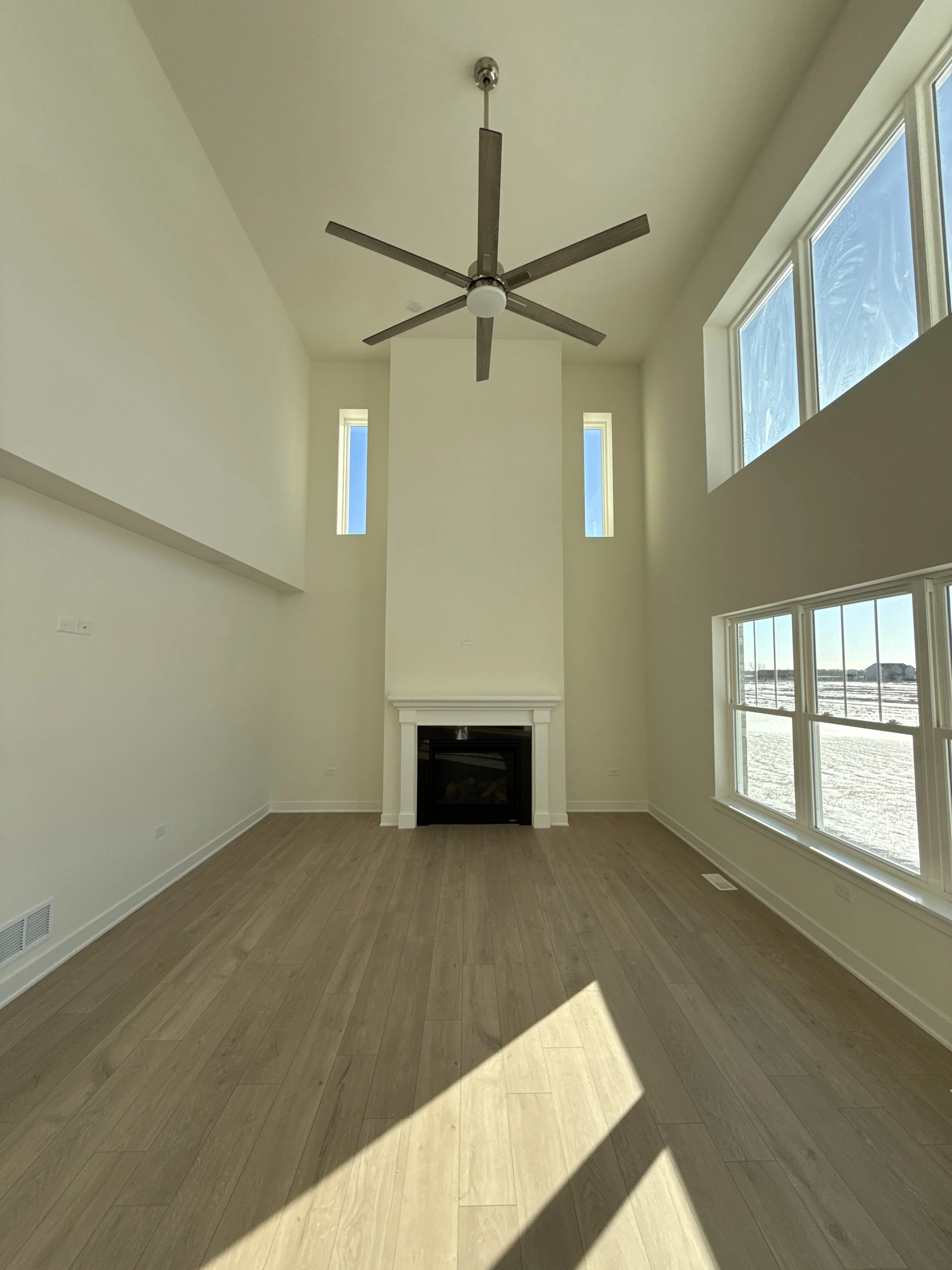 Empty living room with tall ceiling, large windows on the right, a modern ceiling fan, a fireplace with a white mantel, and wood flooring.
