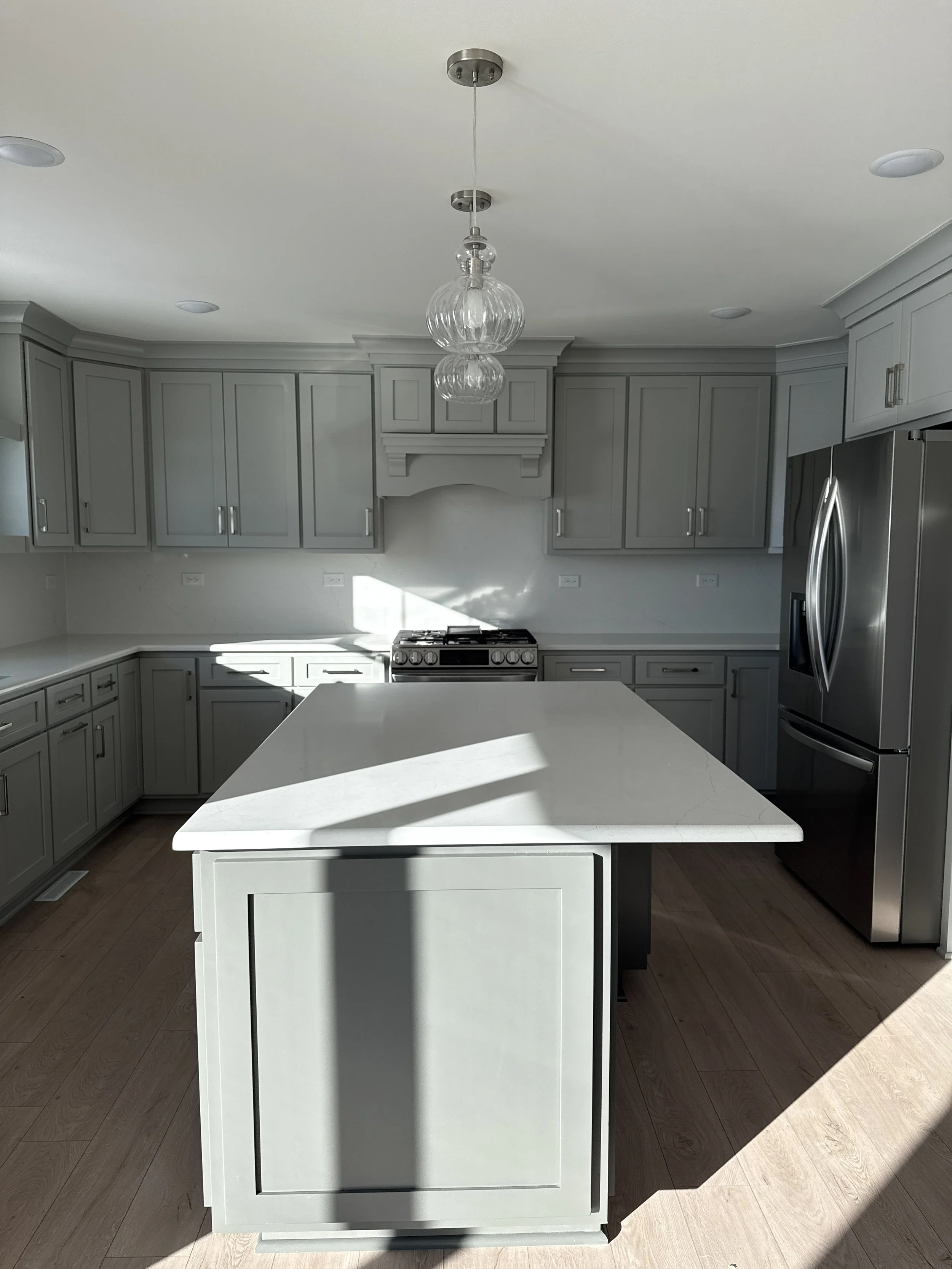 Bright, modern kitchen with light gray cabinets, white countertop island, stainless steel refrigerator, and a kitchen stove. Sunlight casts shadows through windows.