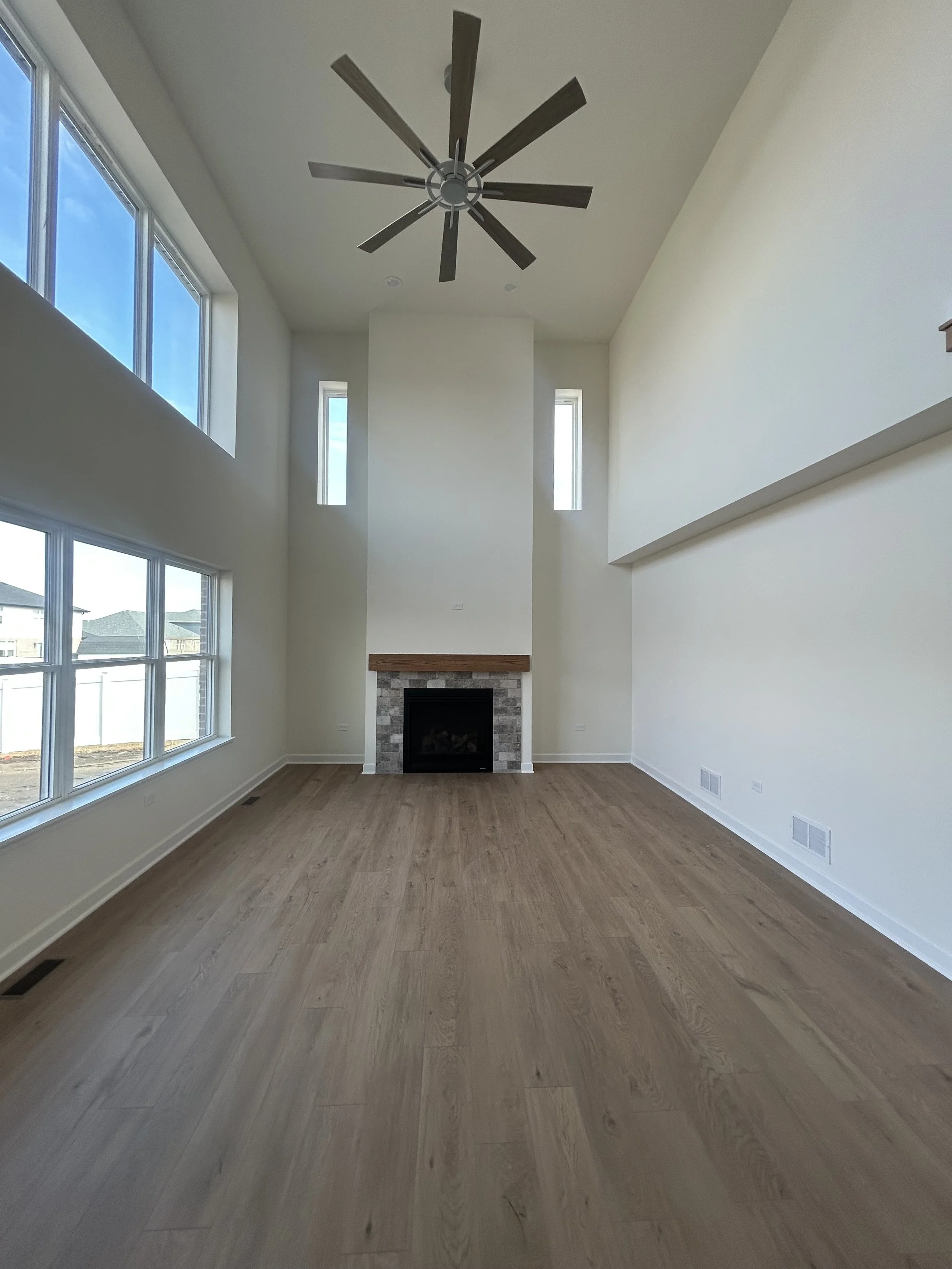 Empty living room with large windows, a fireplace with a stone surround and wooden mantle, a high ceiling with a modern ceiling fan, and light-colored hardwood floors.