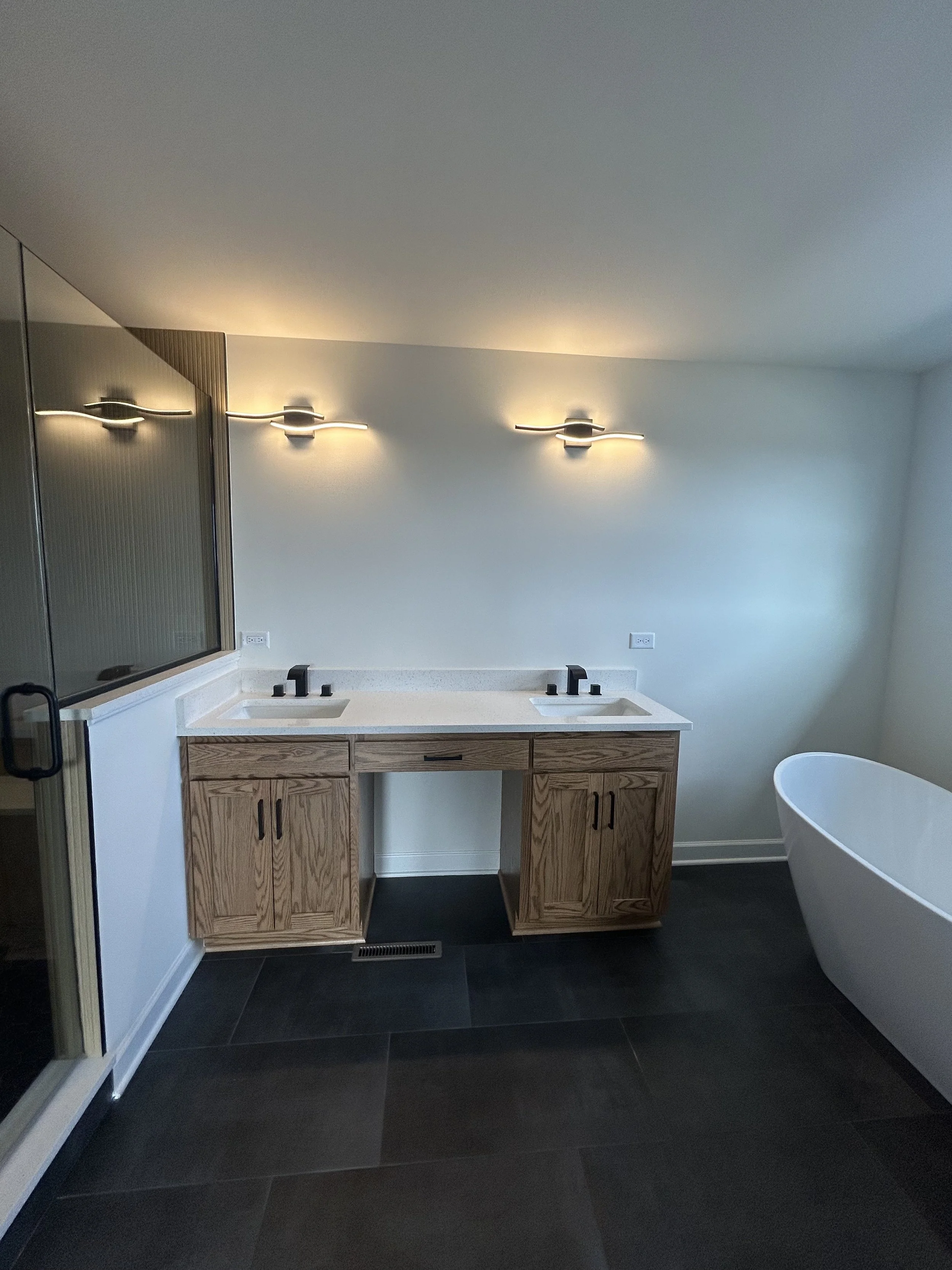 Modern bathroom with double vanity sinks, wooden cabinets, black faucets, wall-mounted lights, a bathtub, a glass shower door, and dark floor tiles.