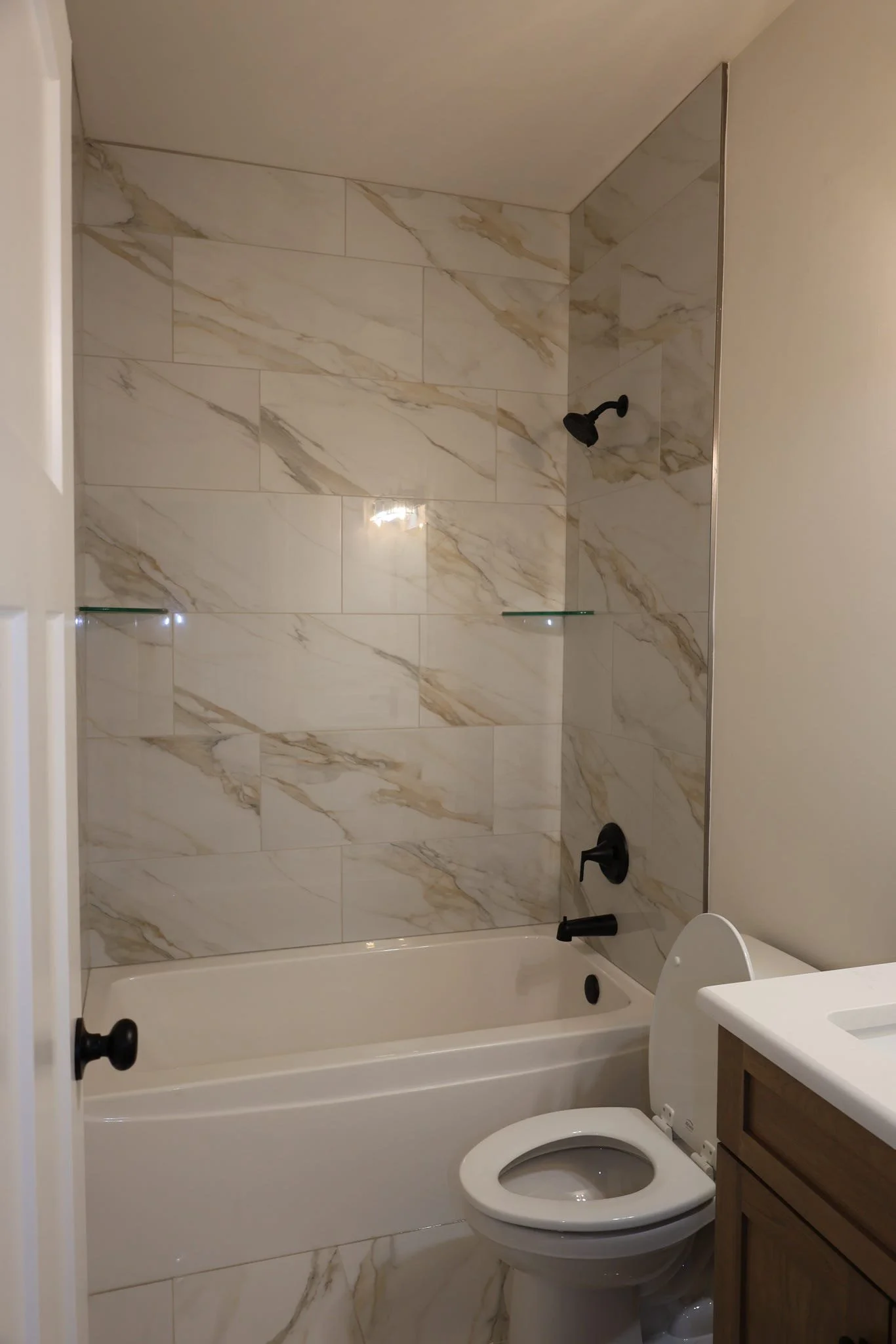 A bathroom with a bathtub, a marble tiled wall, a black shower head and faucet, a toilet with a closed lid, and a wooden vanity with a white sink.