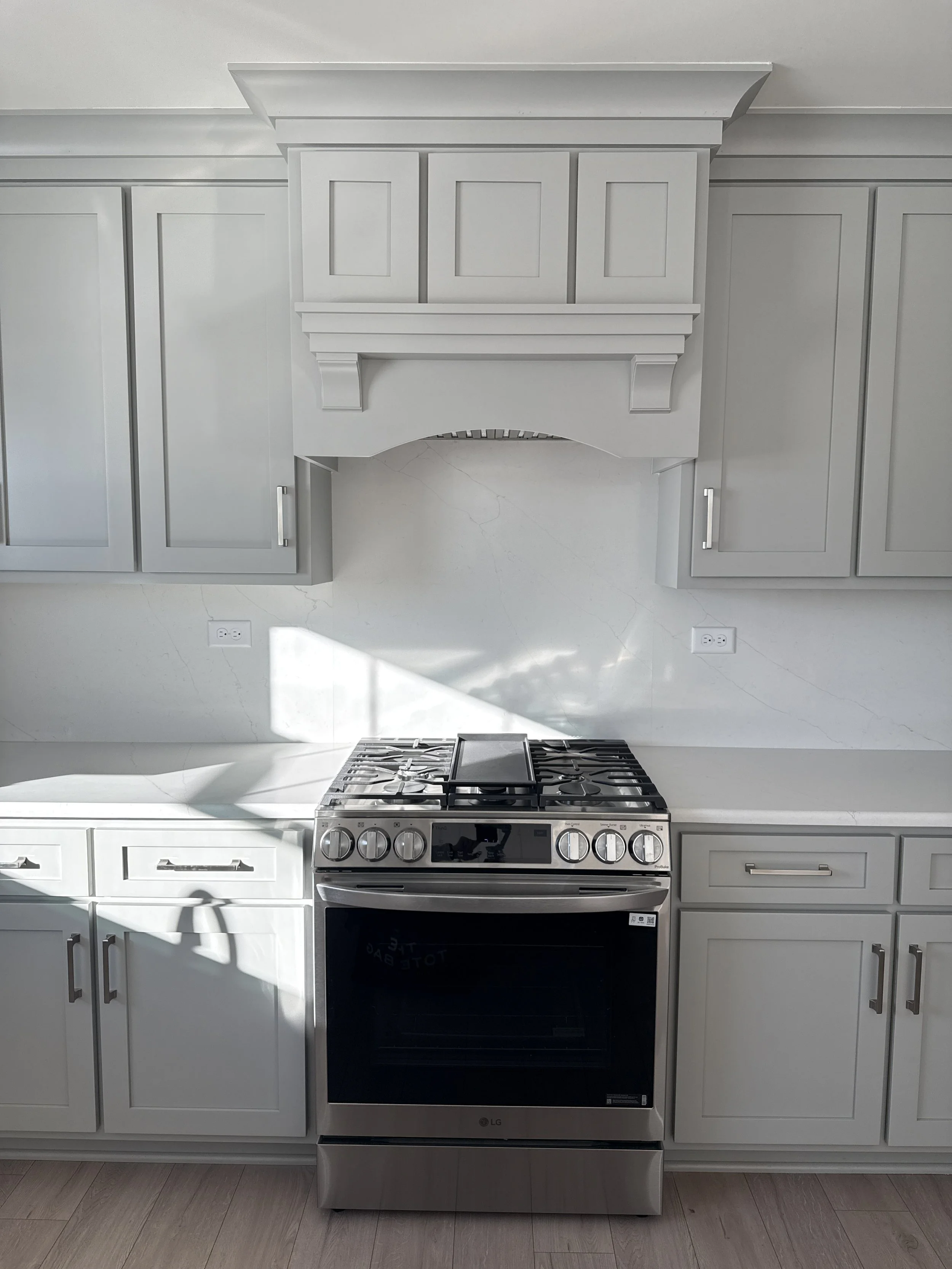 White kitchen with gray cabinets, stove, and decorative range hood. Natural light casting shadows on the countertop and wall.