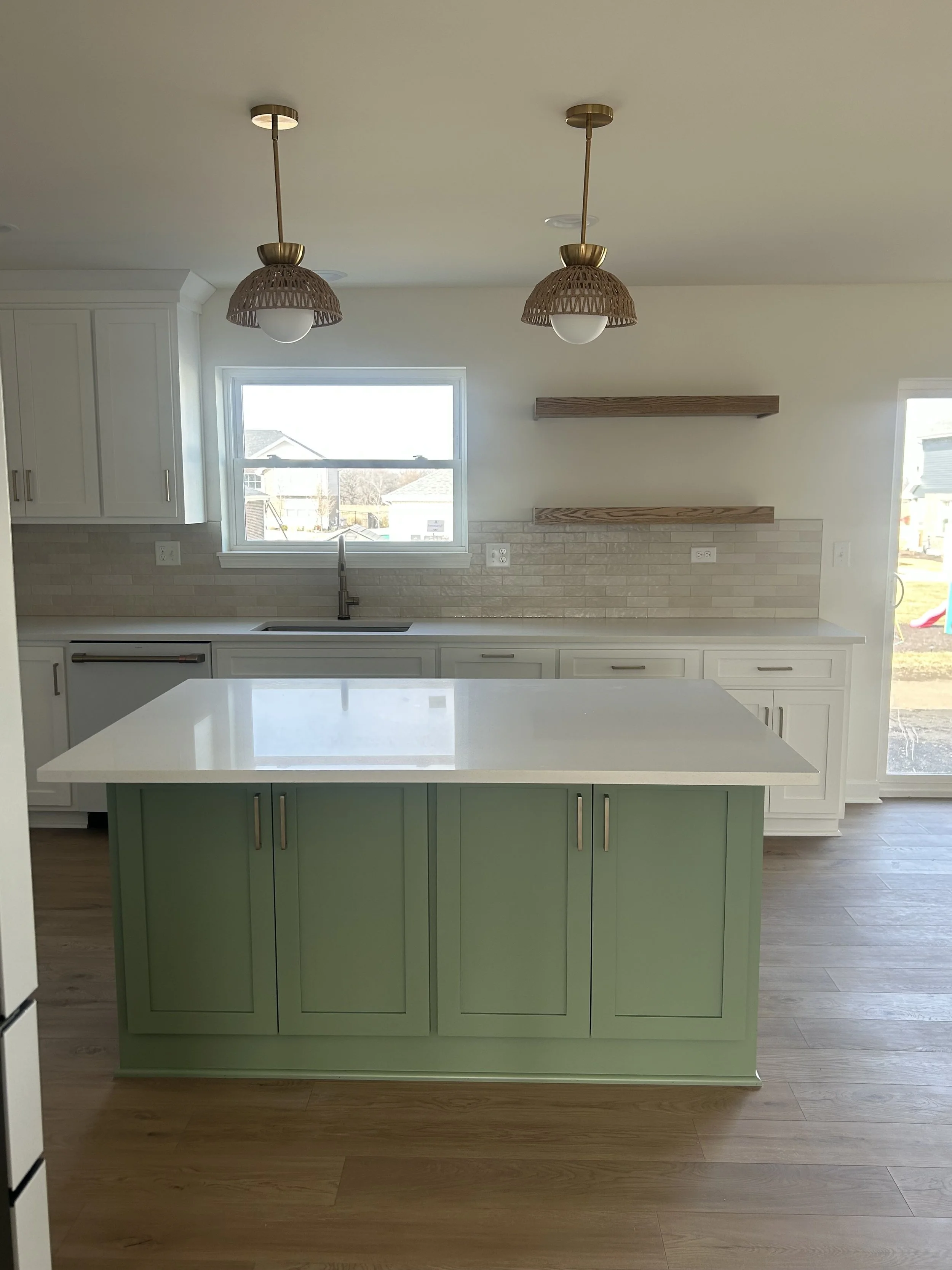 Modern kitchen with a central green island, white countertops, white cabinetry, and beige brick backsplash. Two pendant lights with woven shades hang from the ceiling. A window above the sink allows natural light, and there are two wooden floating sh