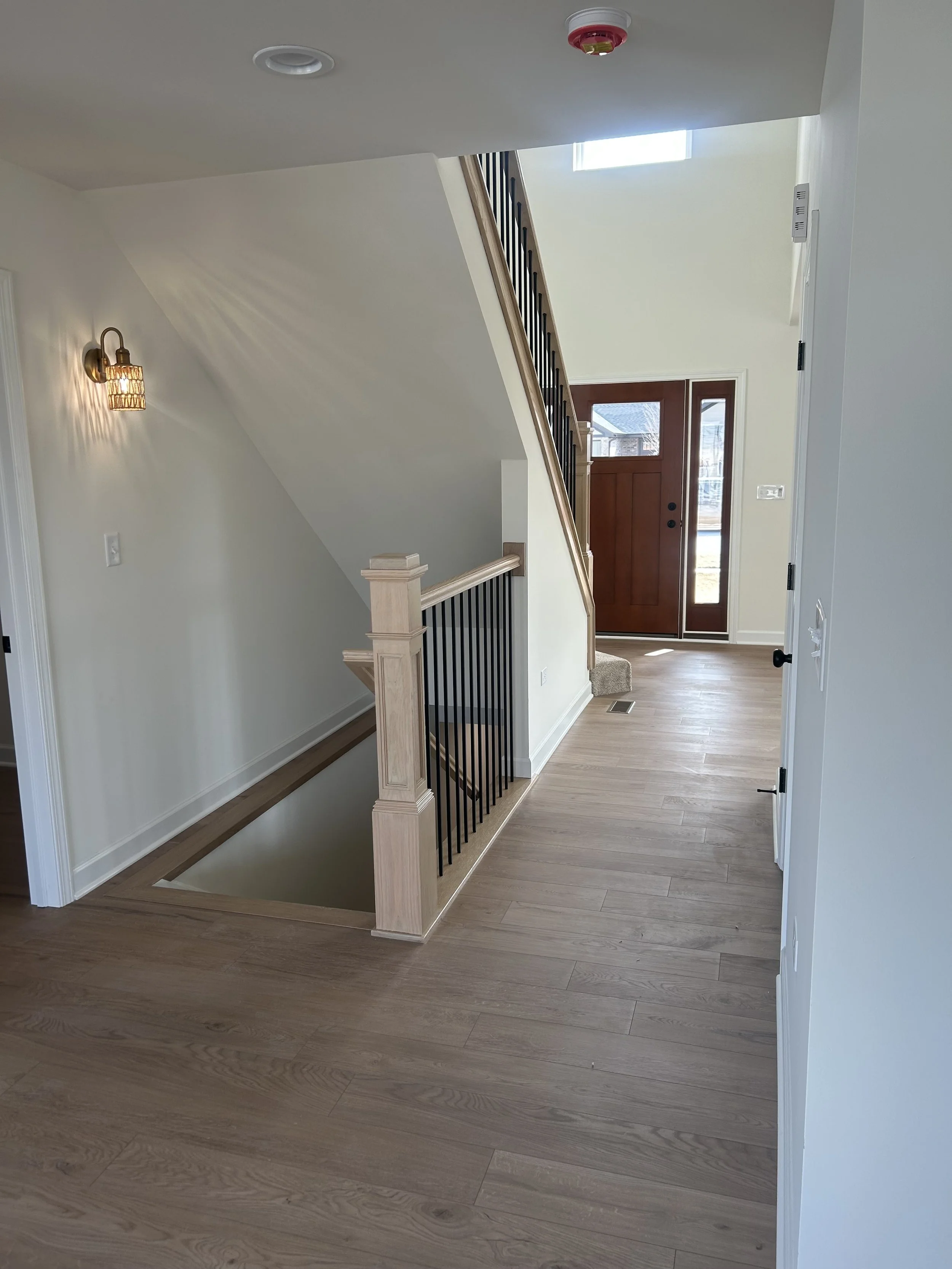 Entryway of a house with front door, staircase, hardwood floors, wall-mounted light fixture, and a window letting in natural light.