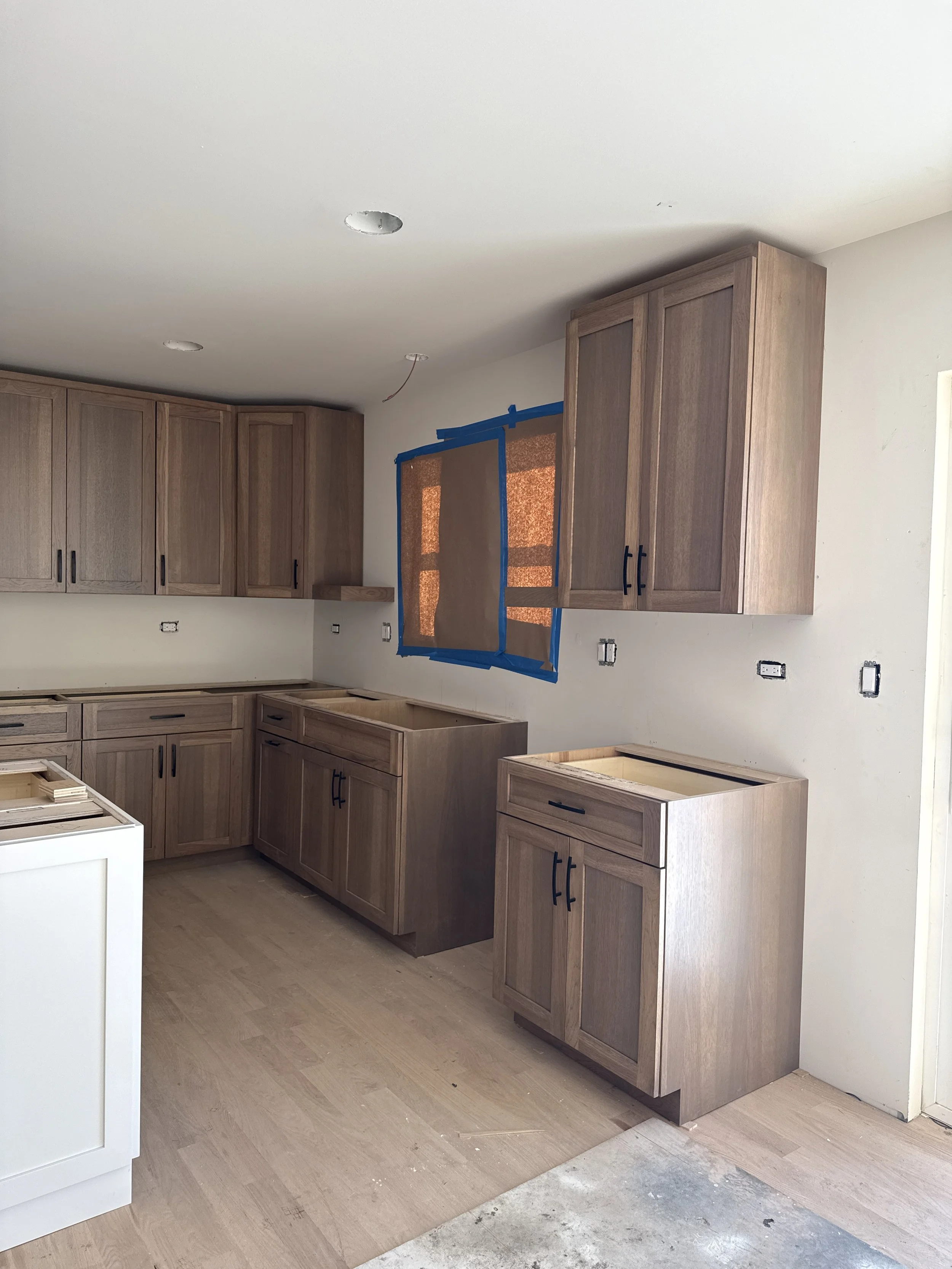 Kitchen cabinets being installed during renovation, with a window covered in brown paper and blue painter's tape, and construction materials on the floor.