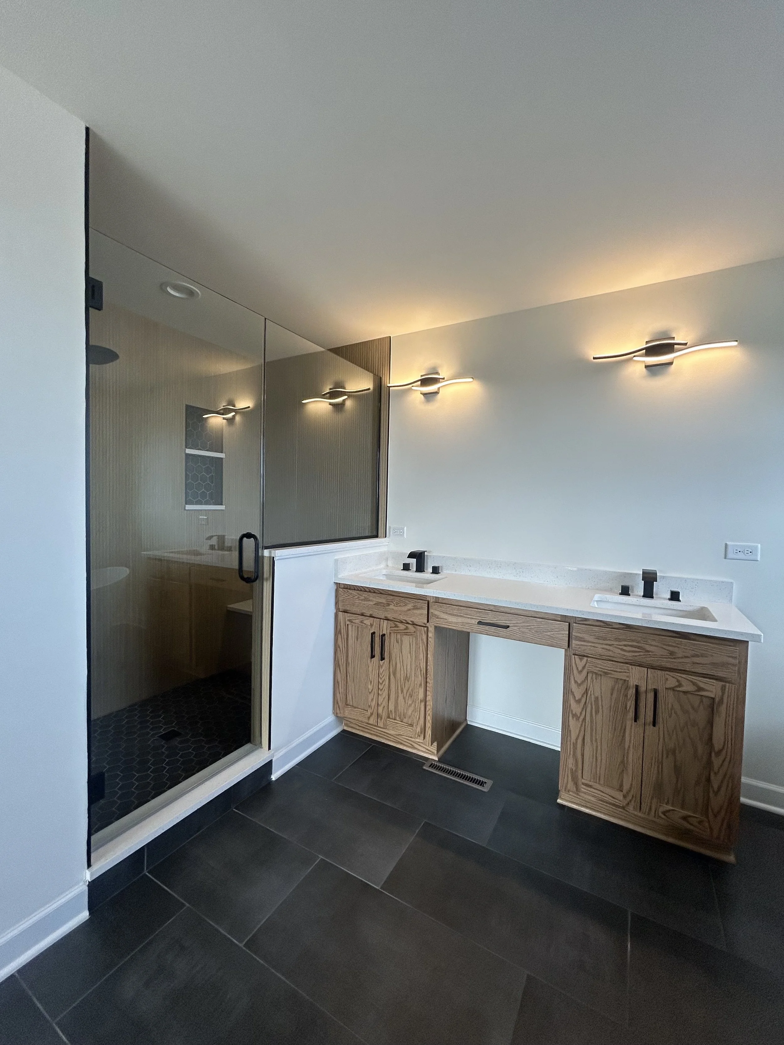 Modern bathroom with a double vanity featuring wooden cabinets, a white countertop, and black fixtures, next to a glass-enclosed shower with a black hexagonal tile floor and a dark sliding door.