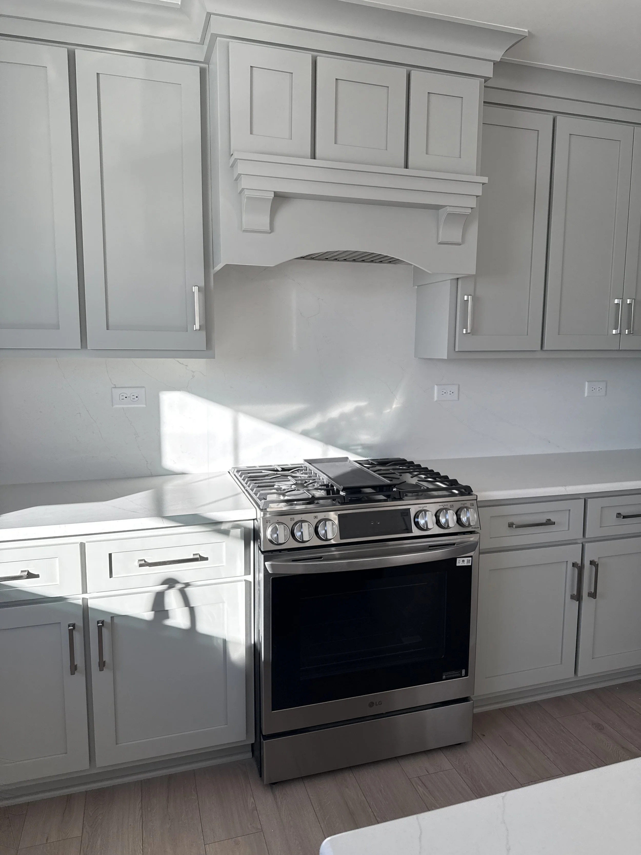 White kitchen with gray cabinets and stainless steel oven, with sunlight casting shadows on the wall.