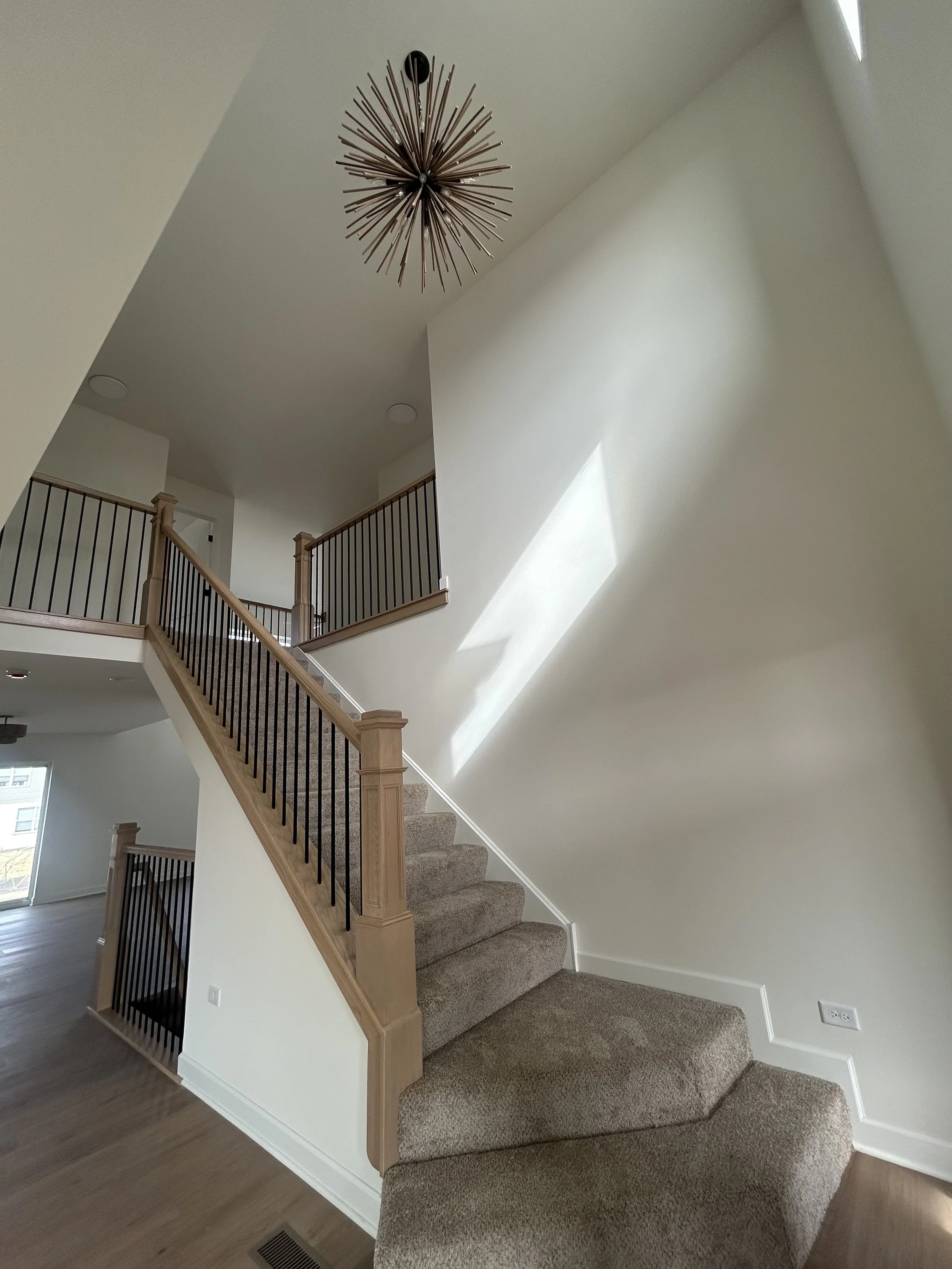 Interior view of a residential staircase covered in beige carpet, with wooden handrails and black spindles, leading to the upper floor, with natural light coming through windows.