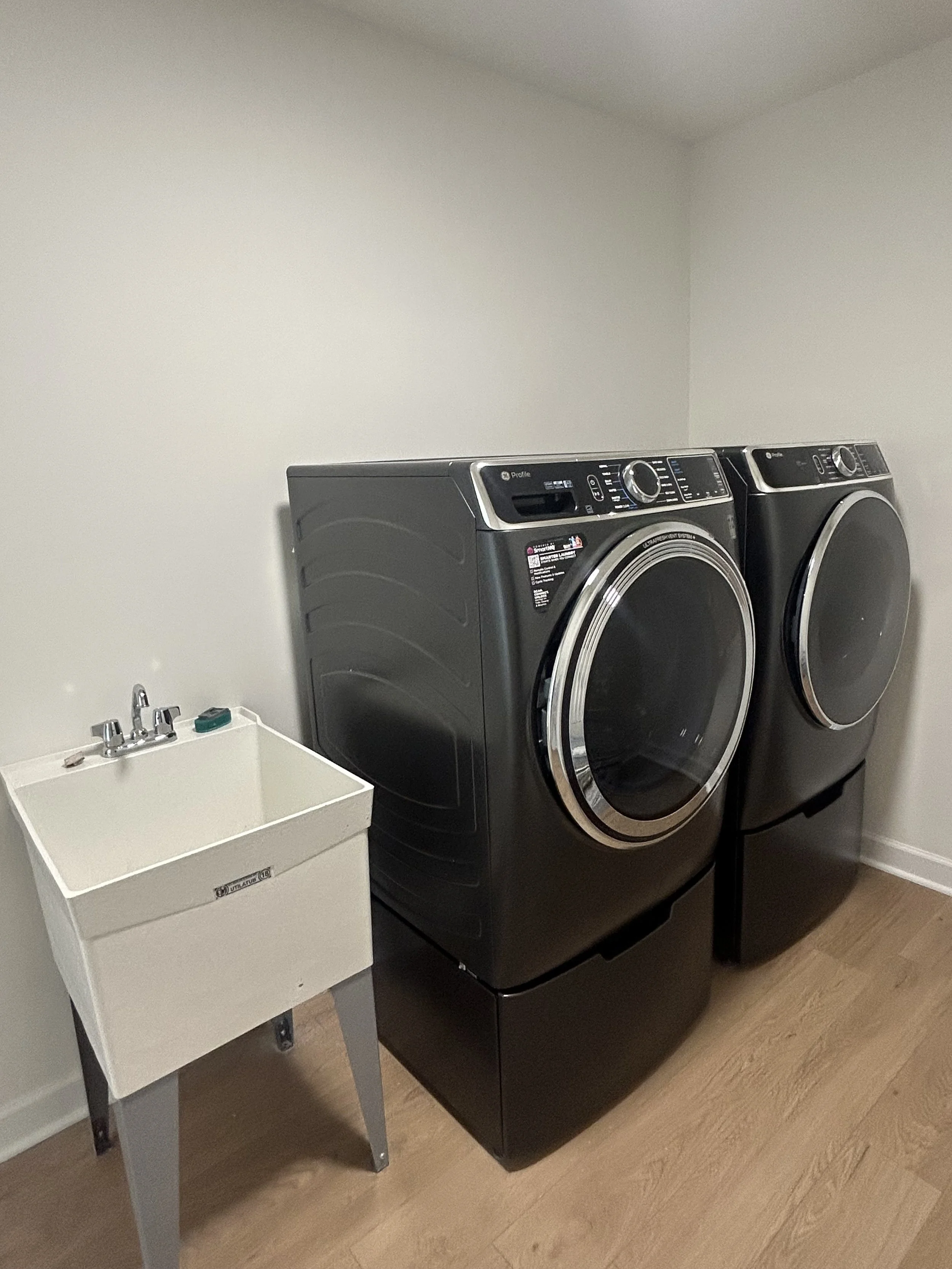 A laundry room with a black front-loading washer and dryer next to each other, and a white utility sink to the left.