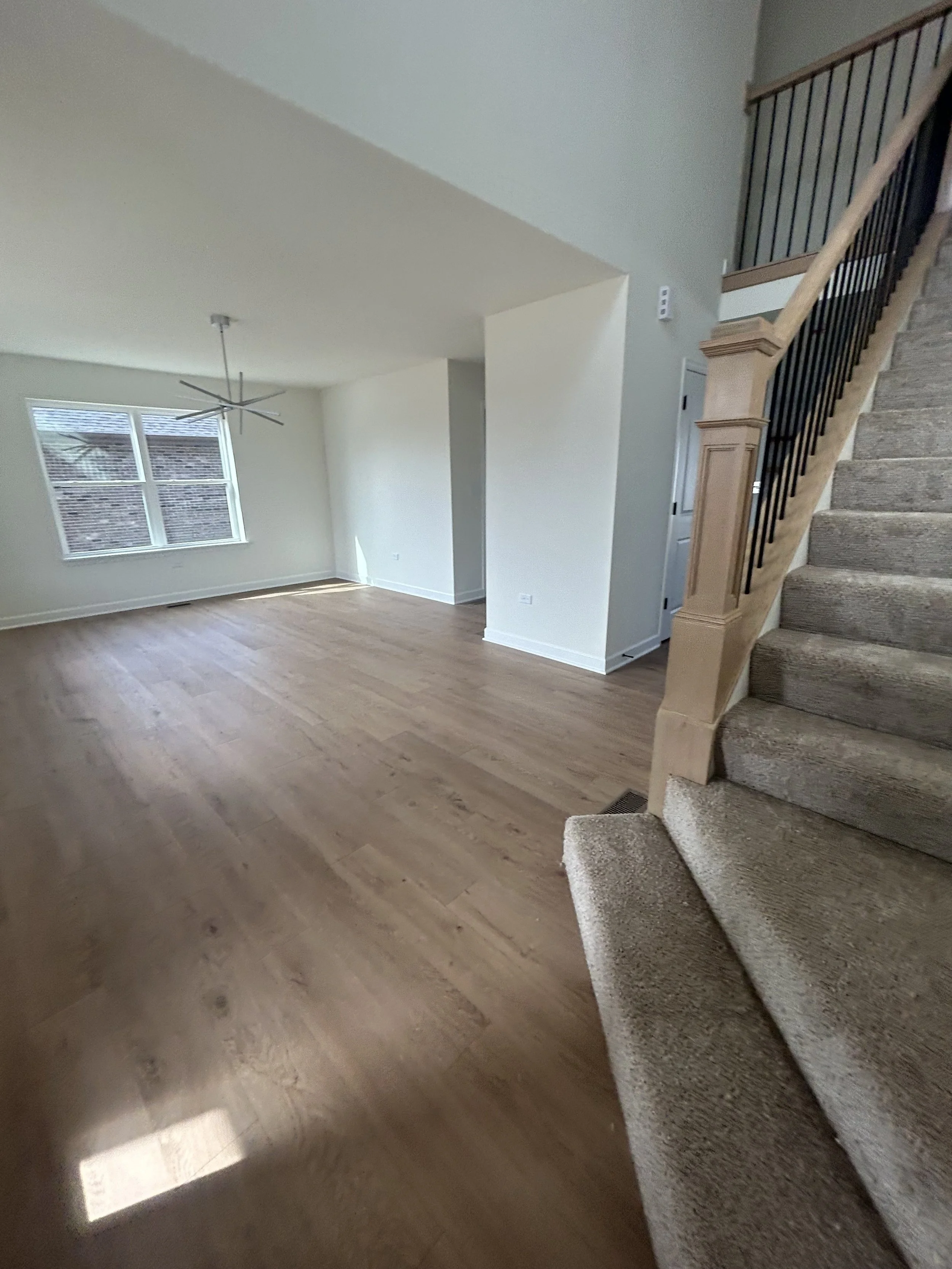 Interior of an unfurnished living room with light wood flooring, a staircase with carpeted steps and wooden railing, a large window with blinds, and a modern ceiling fan.