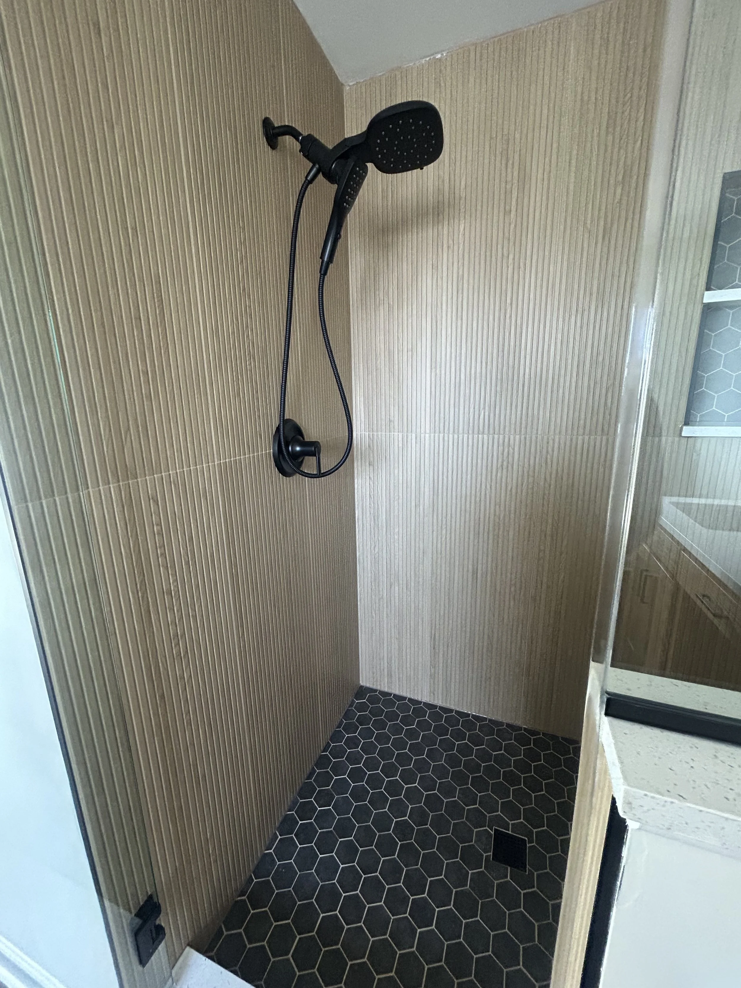 Shower area with a black handheld showerhead mounted on a light brown textured wall, black hexagonal tile floor, and a small floor drain.
