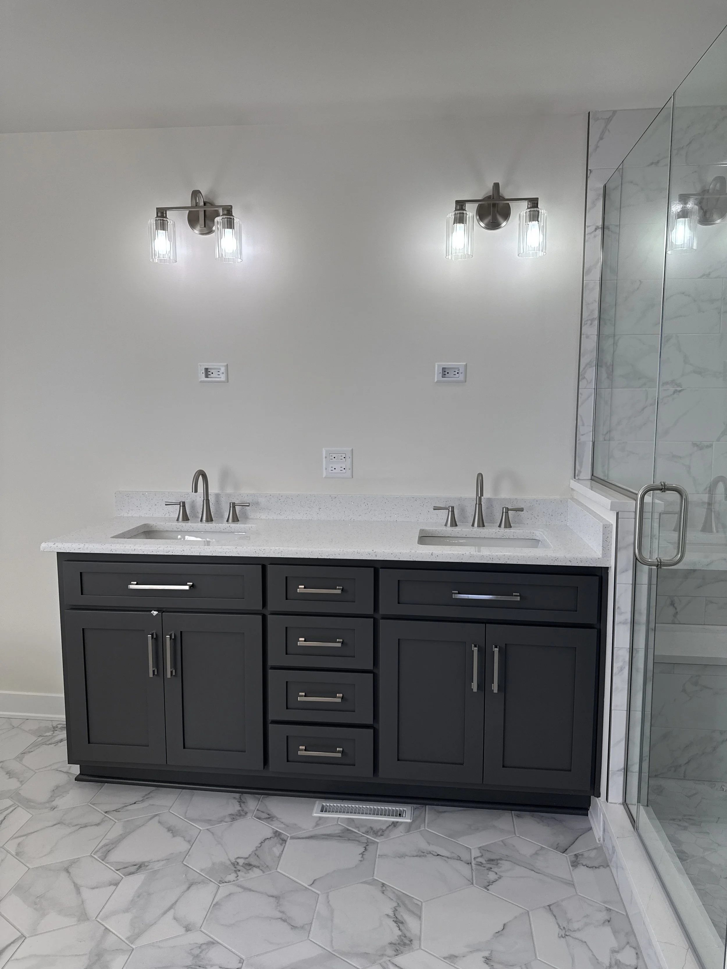 Bathroom vanity with black cabinetry, white speckled countertops, two sinks, wall-mounted faucets, and modern lighting fixtures above, next to a glass shower enclosure with marble tiles and a chrome handle, marble tile flooring.