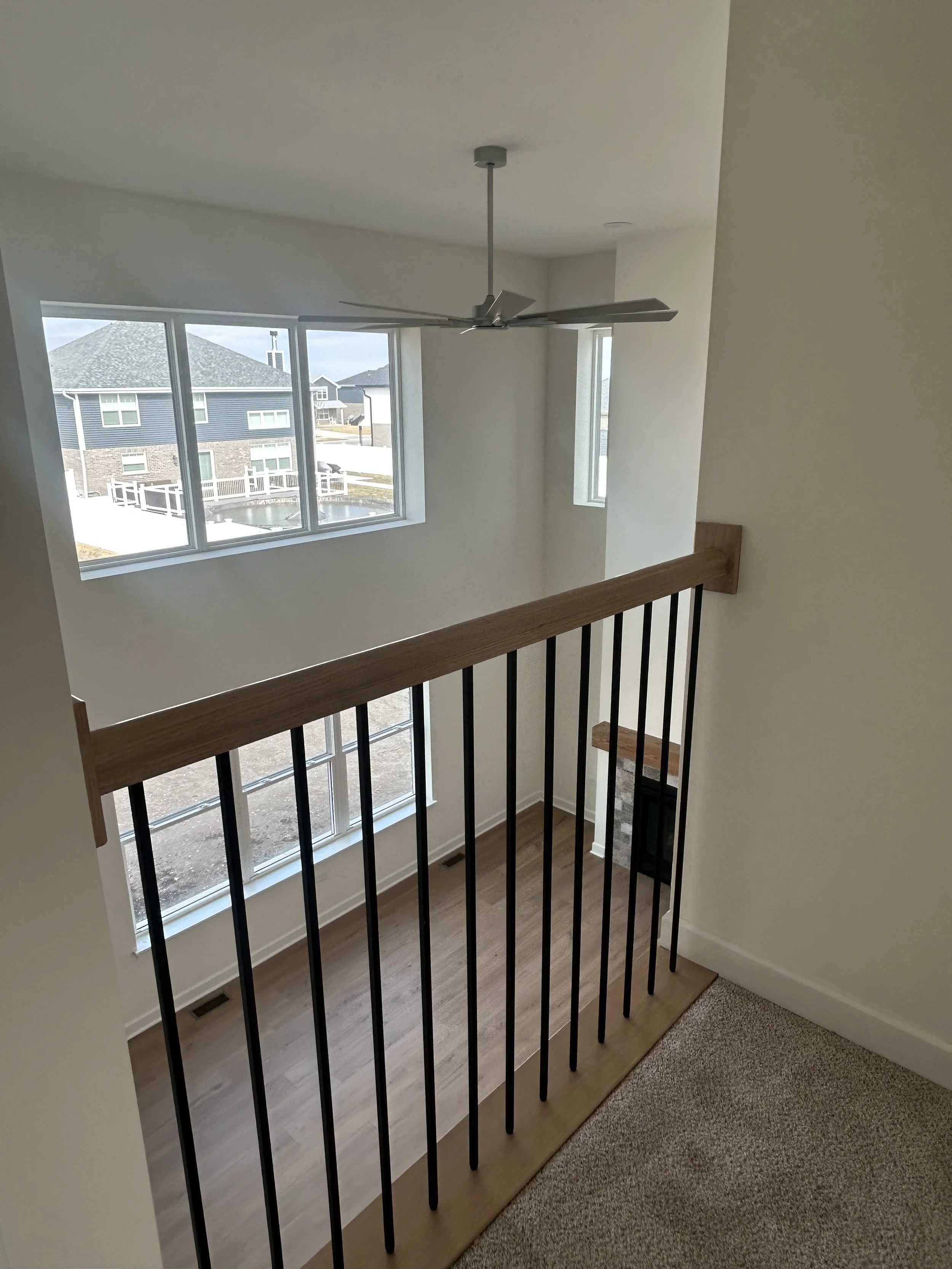 View from an upstairs landing of a house interior with large windows, a ceiling fan, and a fireplace below. The balcony has a wooden and black metal railing.