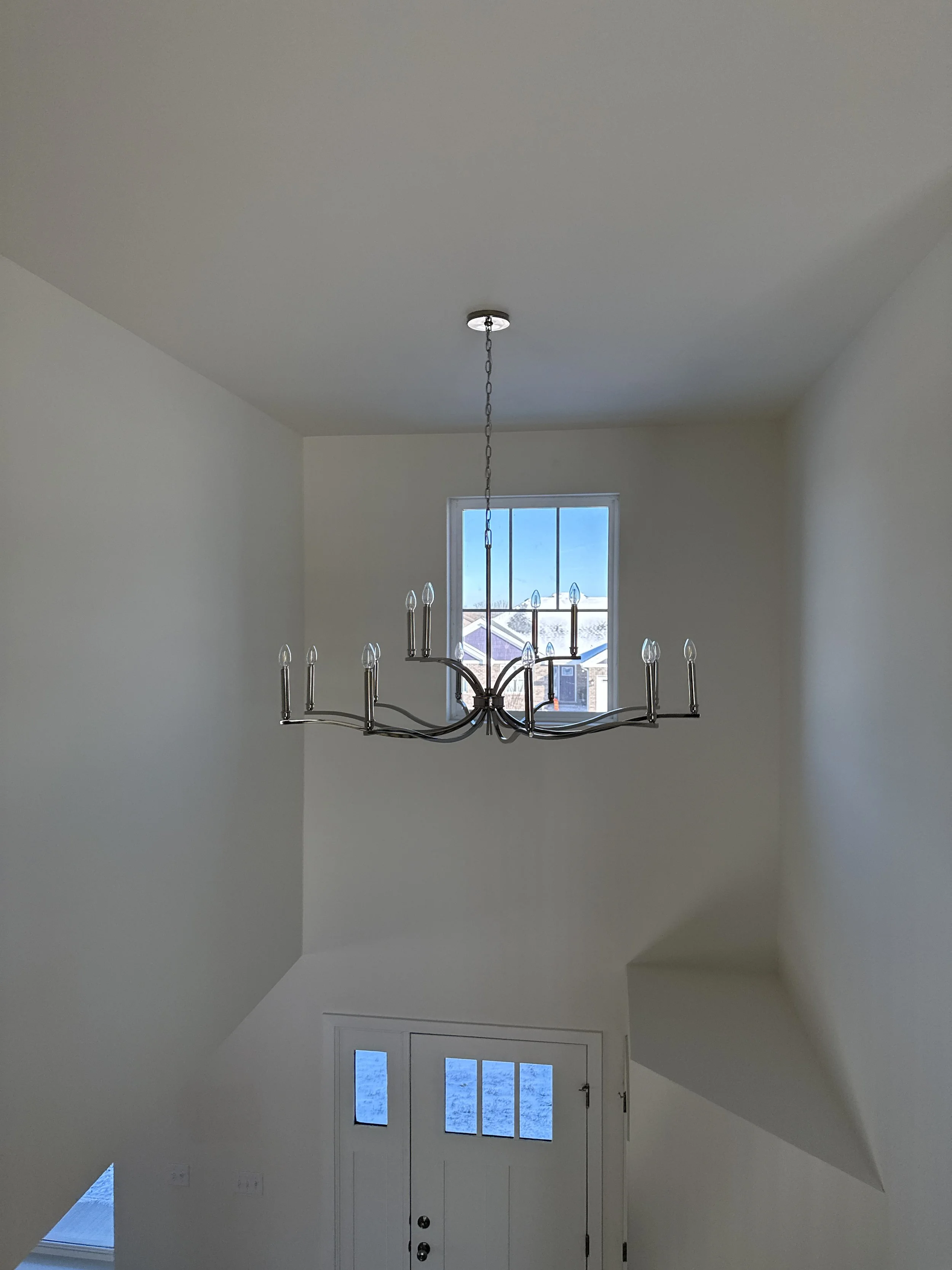 Empty room with a metal chandelier hanging from the ceiling, a window, and a white front door beneath the window.
