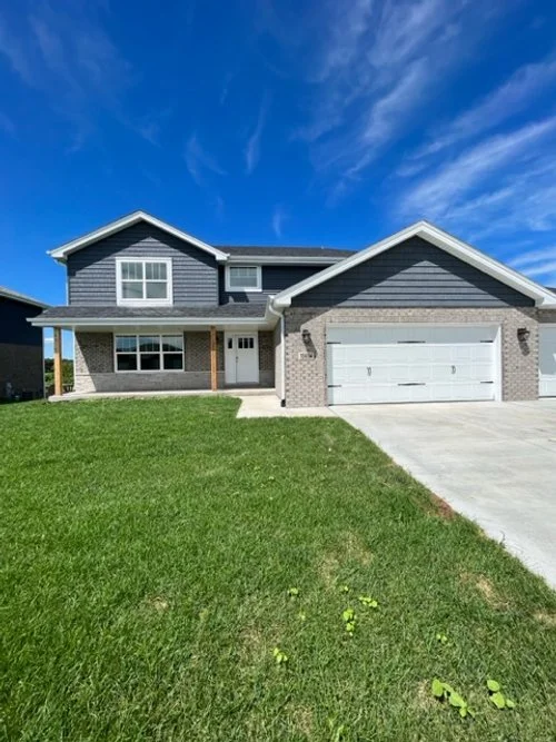 Two-story house with gray siding, brick accents, a white garage door, and a front lawn under a blue sky with wispy clouds.
