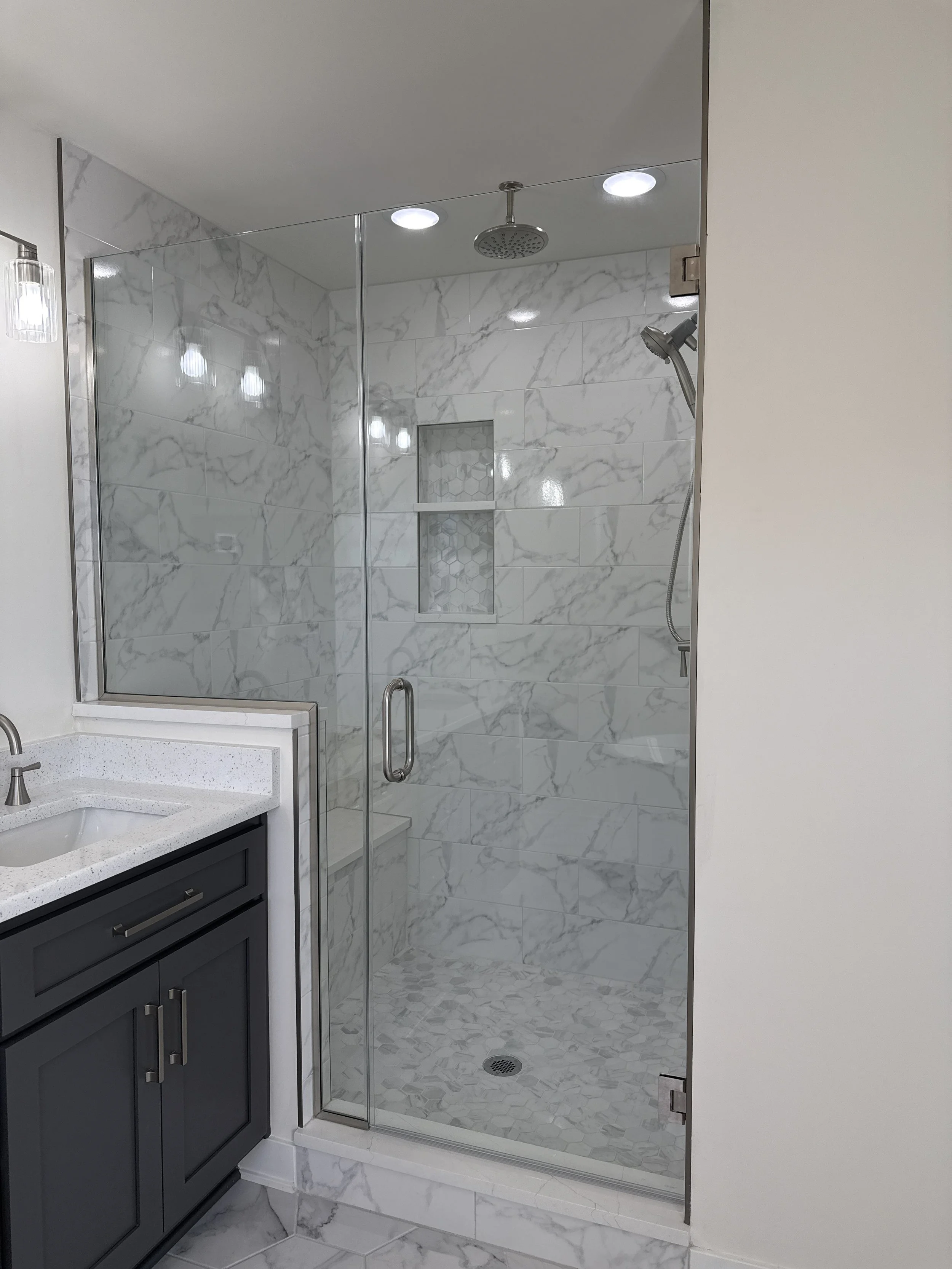 A modern bathroom with a glass-enclosed shower featuring marble tiles, a built-in niche, and a rain showerhead. Part of a vanity with a dark cabinet and light countertop is visible to the left.