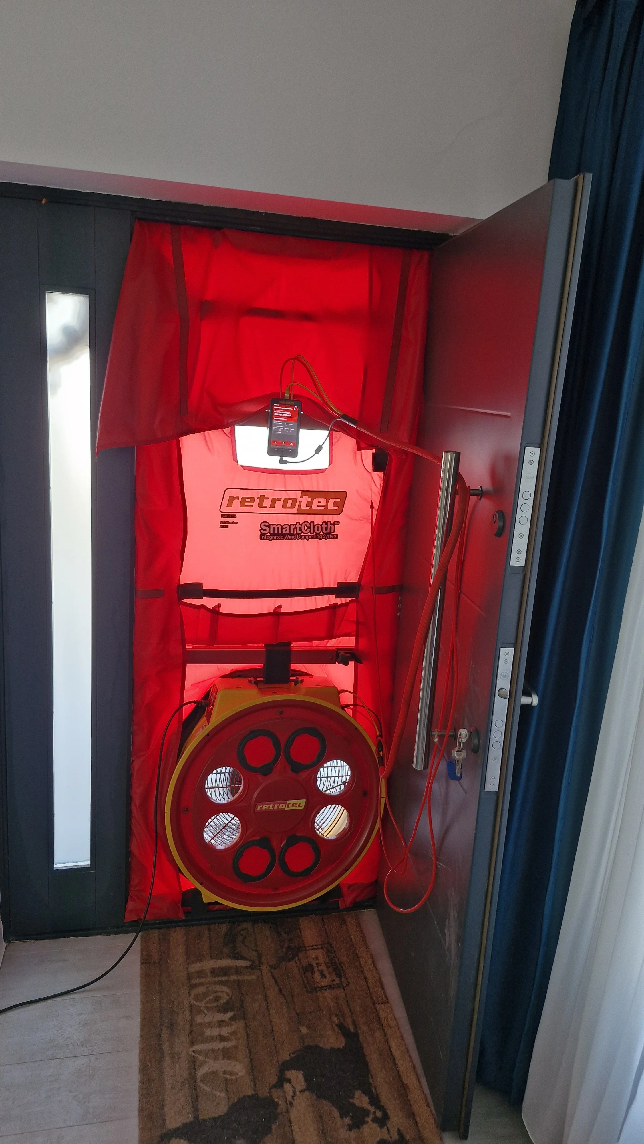 A red retrotec SmartCloth wind testing system setup in a doorway, with various cables and controls.