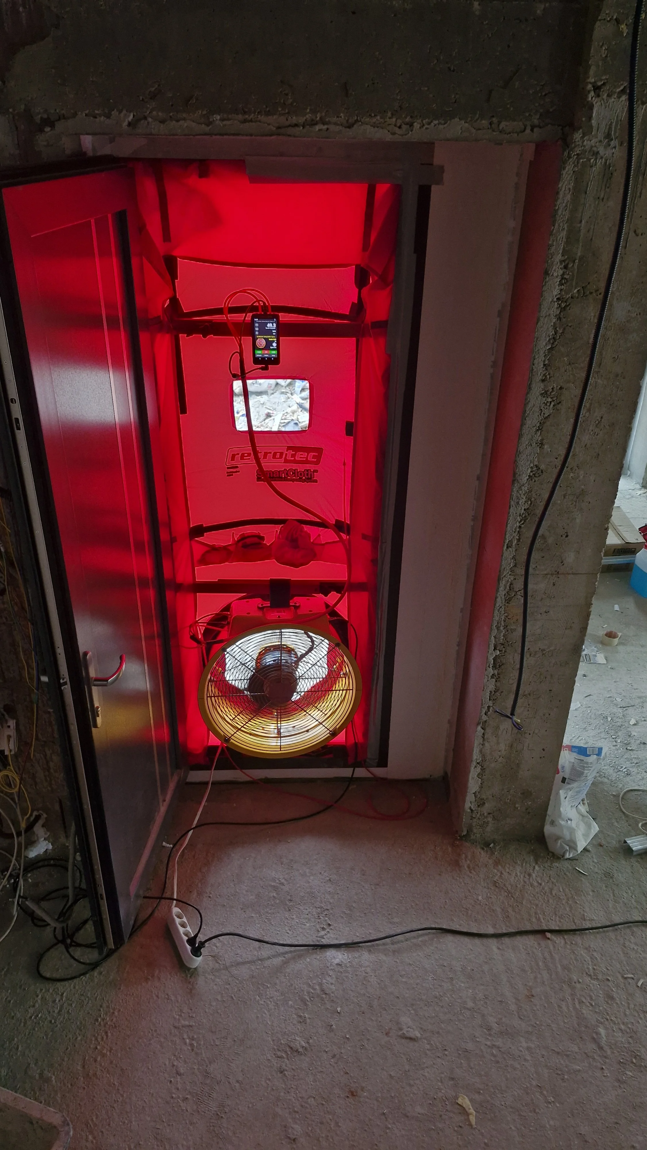 A red light therapy device inside a construction site doorway, with a fan at the bottom, wires, and a control panel.