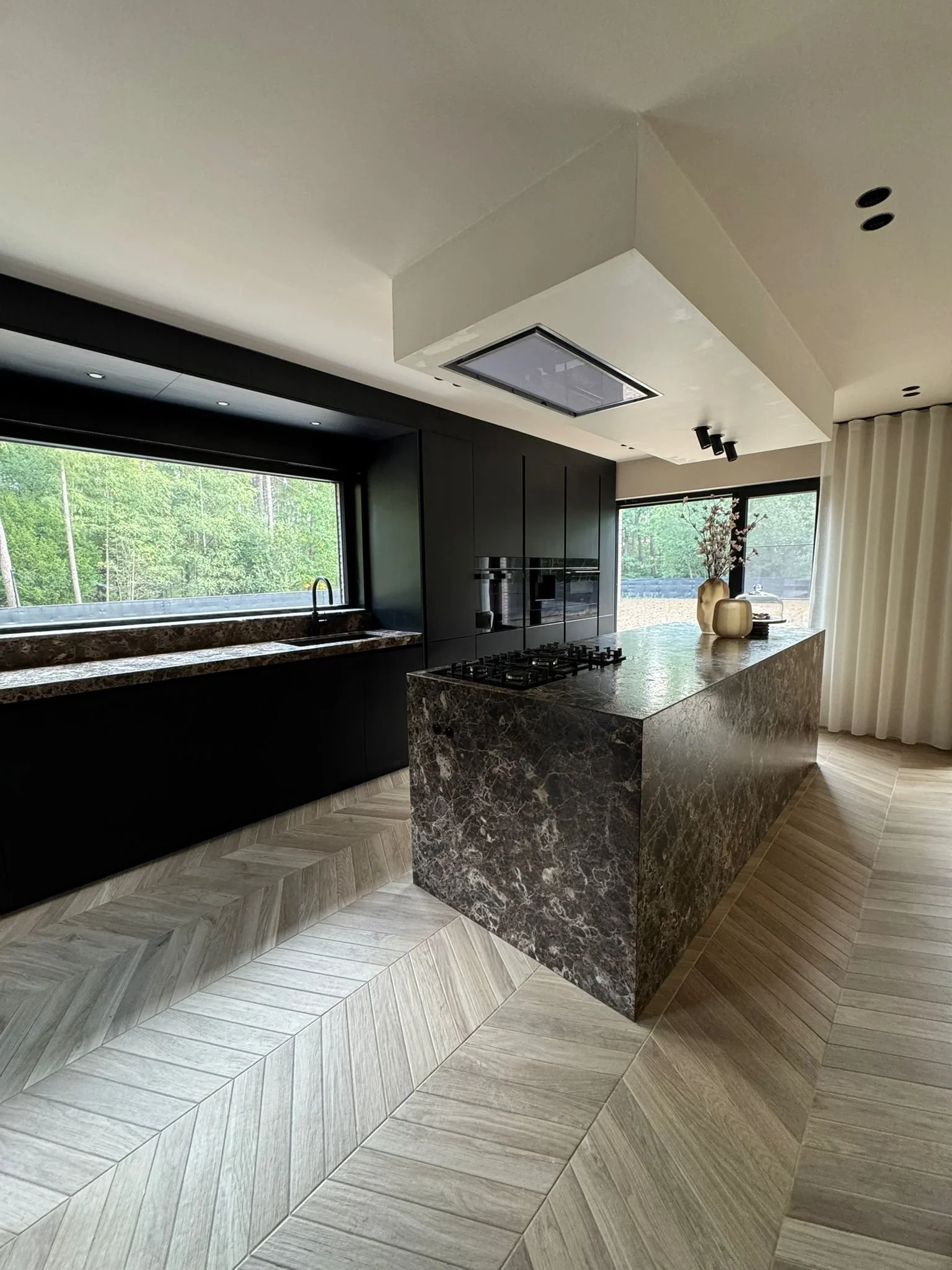 Modern kitchen with black cabinetry, marble countertops, and a marble kitchen island. Large windows with a view of greenery, light wood chevron flooring, and white ceiling with recessed lighting.
