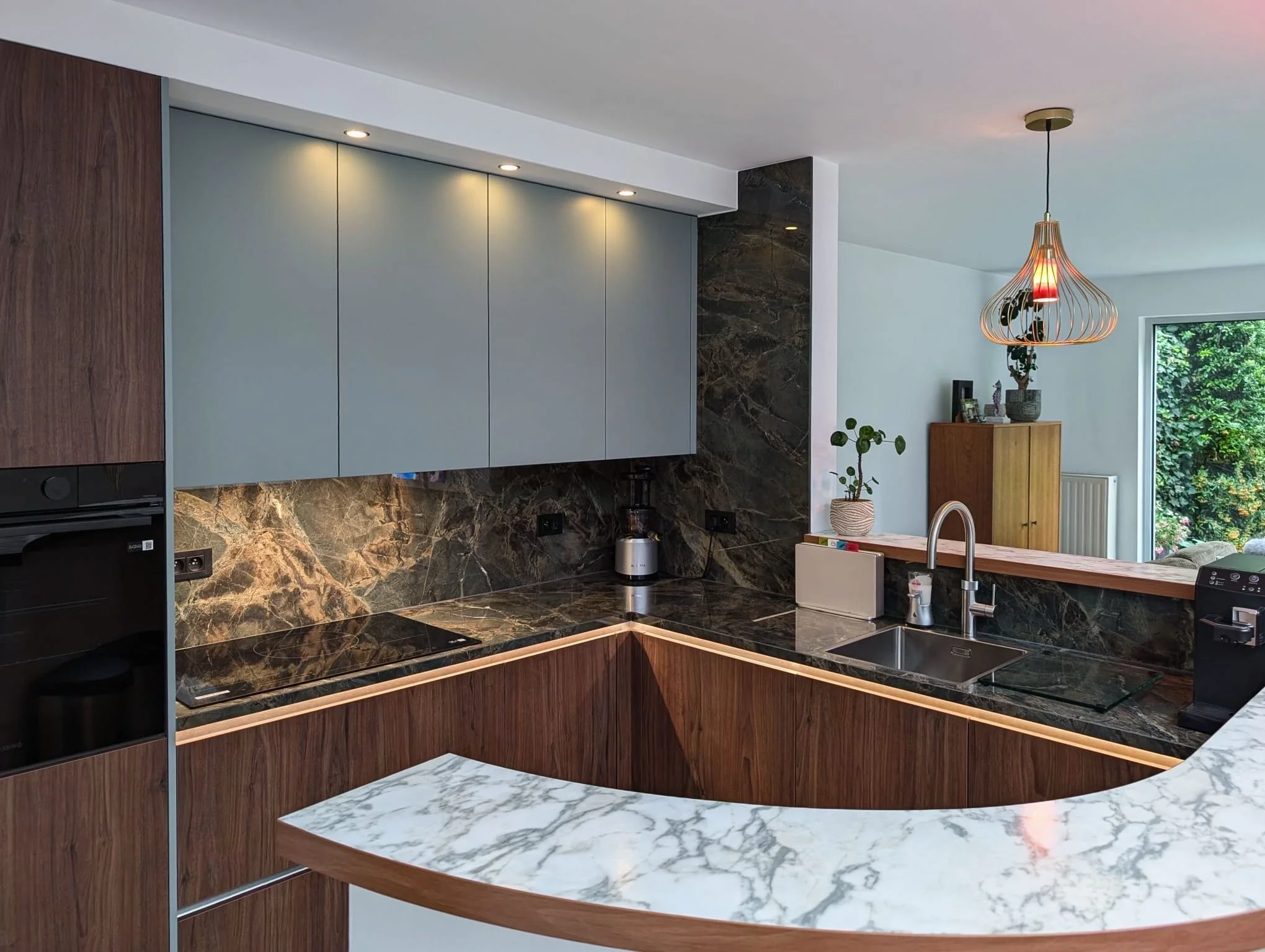 Modern kitchen with dark marble countertops, light grey cabinets, wooden lower cabinets, and a marble breakfast bar, with a view of a bright room with a hanging pendant light and large window showing greenery outside.