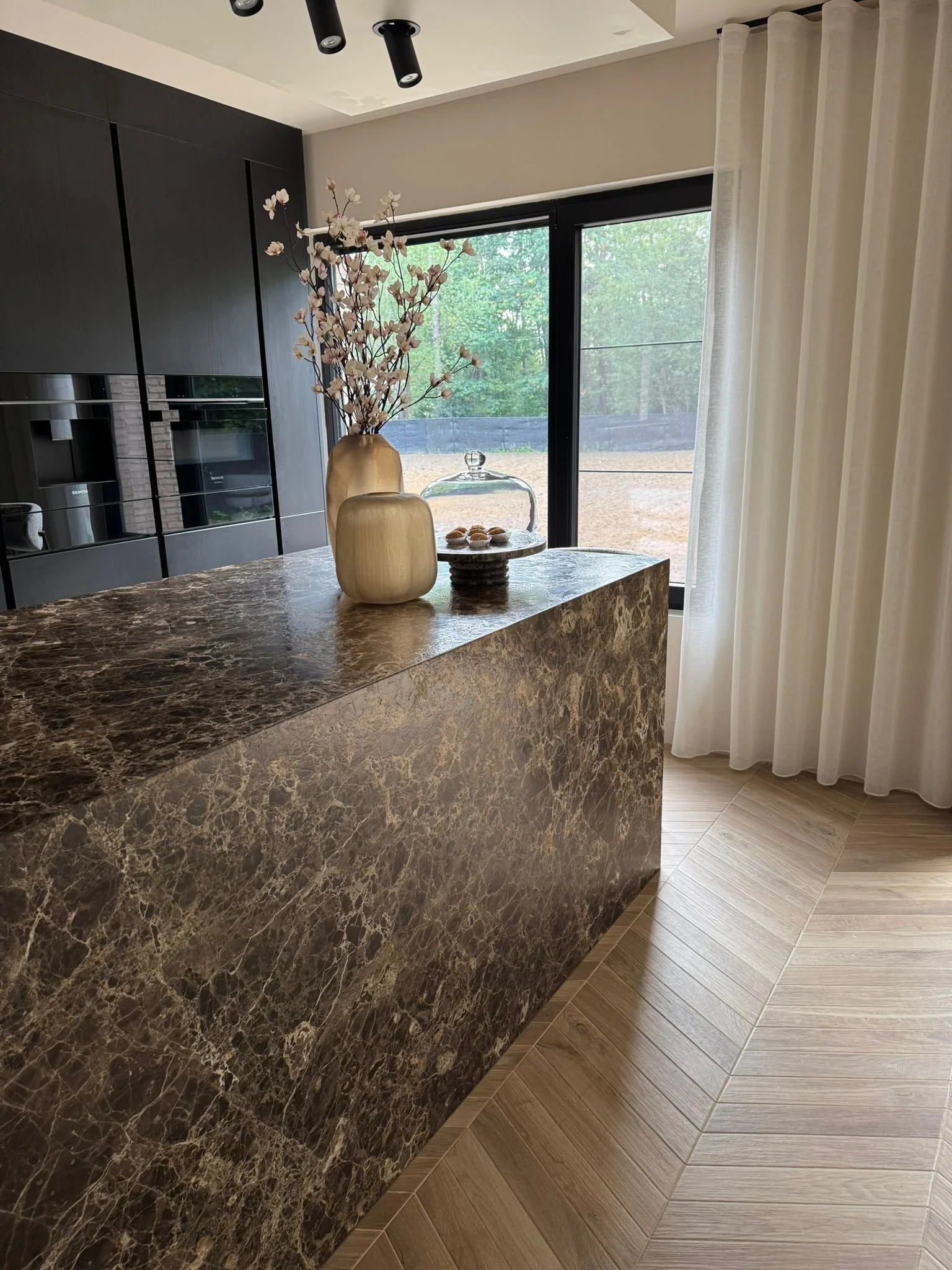 A modern kitchen with a brown marble island, decorative vases with flowers, a cake stand with pastries, and a large window with white curtains.