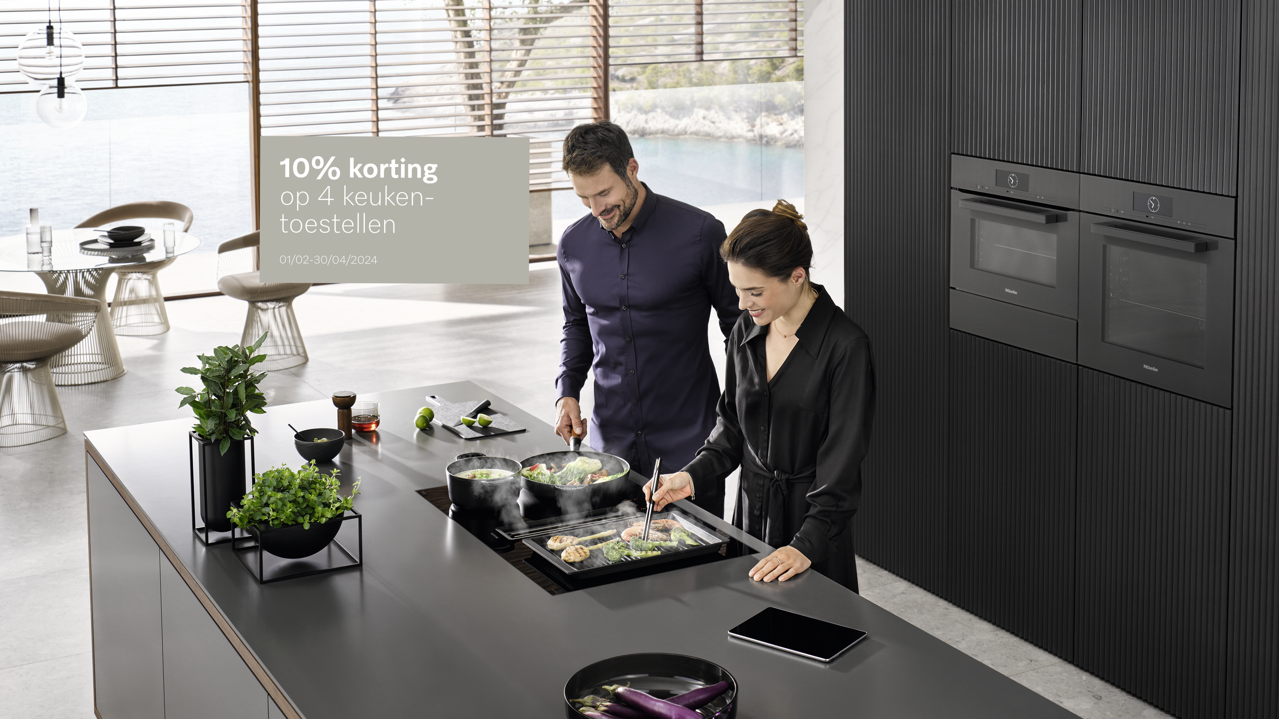 Two people cooking in a modern kitchen with a view of water outside. The man is stirring a pot, and the woman is using tongs to prepare food on an electric stove. There are plants and kitchen items on the island.