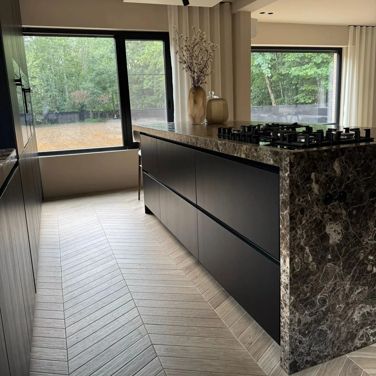 Modern kitchen with black cabinetry, marble countertop, built-in gas stove, large windows overlooking an outdoor yard, decorative vases with flowers on the counter, light-colored wood tilted floor.