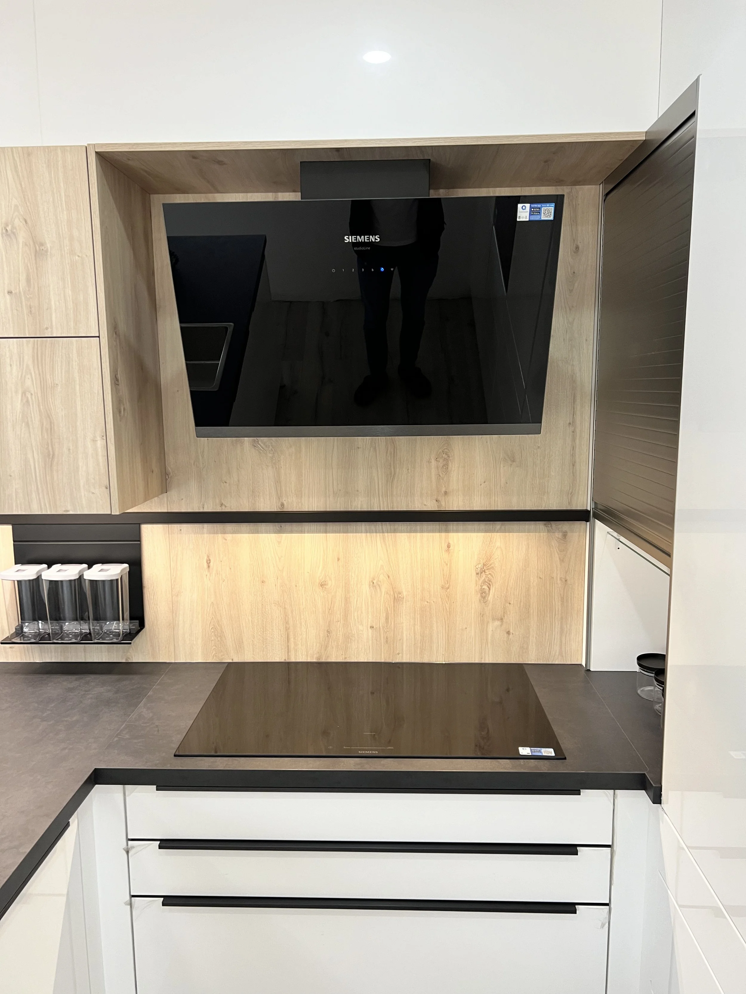 A modern kitchen display with a black Siemens cooktop on a wooden countertop and a black glass wall-mounted oven above, with a reflection of the person taking the photo.
