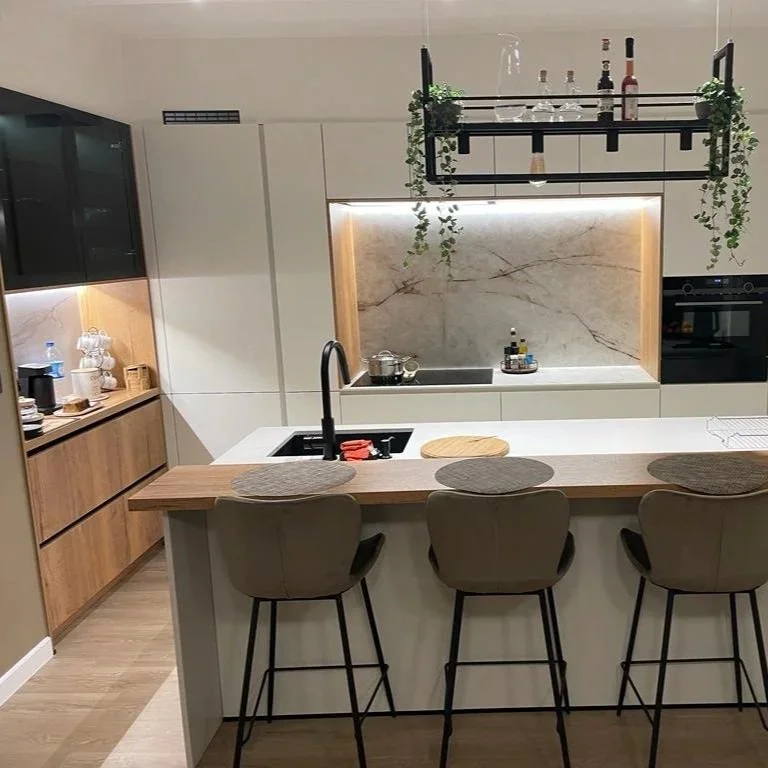 Modern kitchen with a white island, black sink faucet, and three bar stools. Open shelving with glassware above and wooden cabinetry below. Marble backsplash and a hanging pot rack with bottles and greenery.