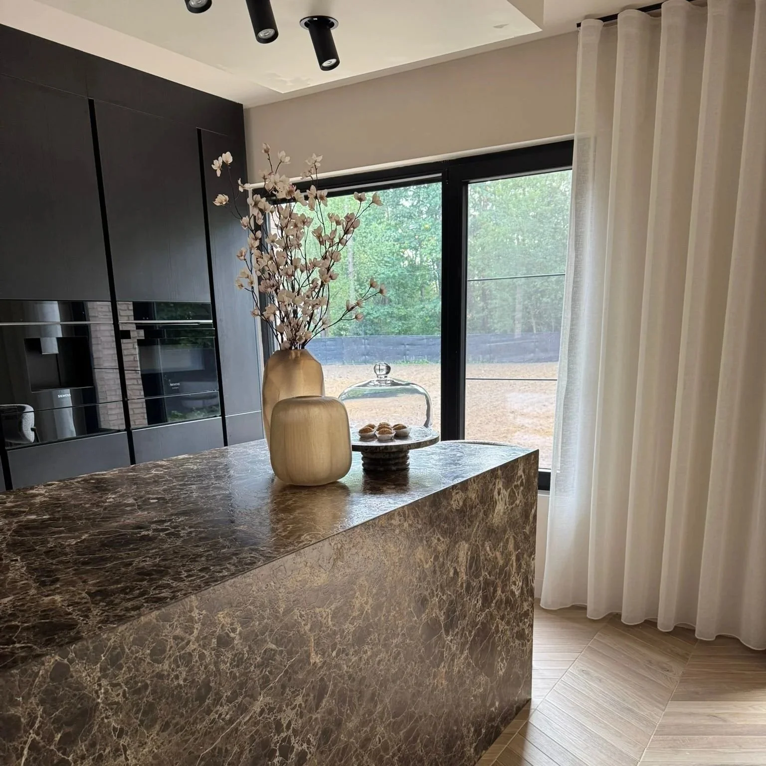 Decorative vases with flowers and pastries on a marble kitchen island in a modern kitchen with large window and white curtains.